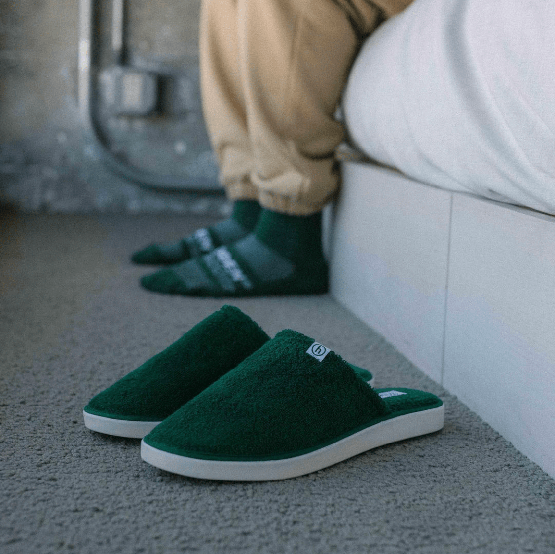 Front three quarter view of green terry cloth hotel slippers with white cushioned sole on carpet by a bed