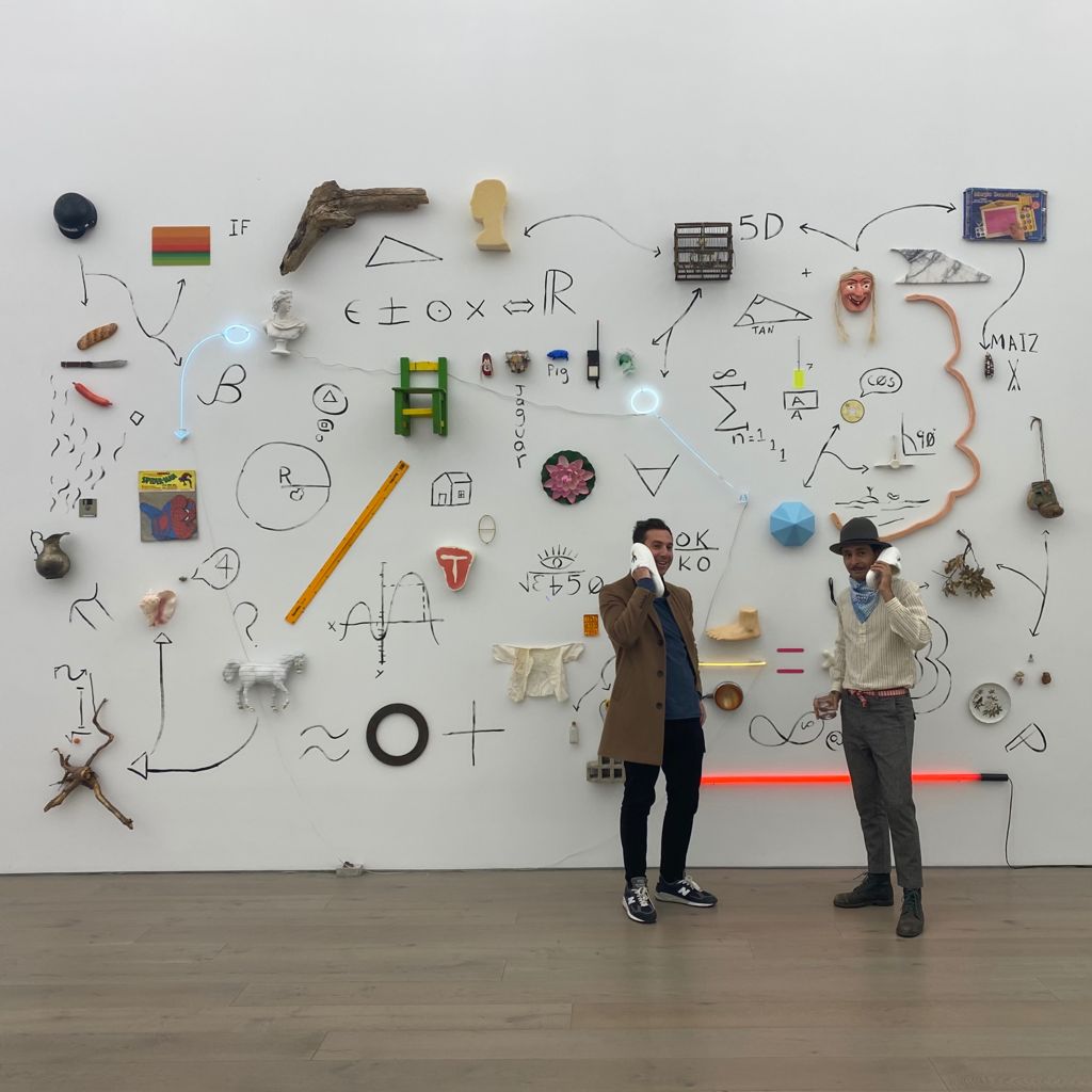 Two men holding phone receivers in front of a large white gallery wall covered with drawings, found objects, and neon accents