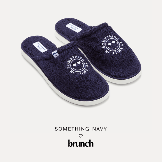 Three-quarter view of navy terry slip-on slippers with embroidered circular Something to smile in logo and white rubber sole on a light background