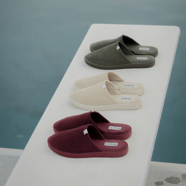 Three-quarter view of three corduroy slippers showing ribbed texture and branded insole on a poolside ledge