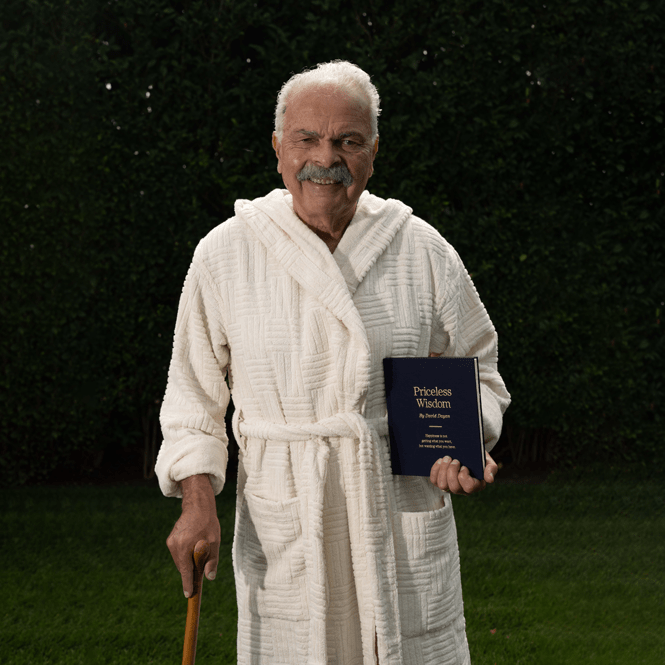 Man standing on grass in a cream robe holding a navy book titled 'Priceless Wisdom' and a wooden cane