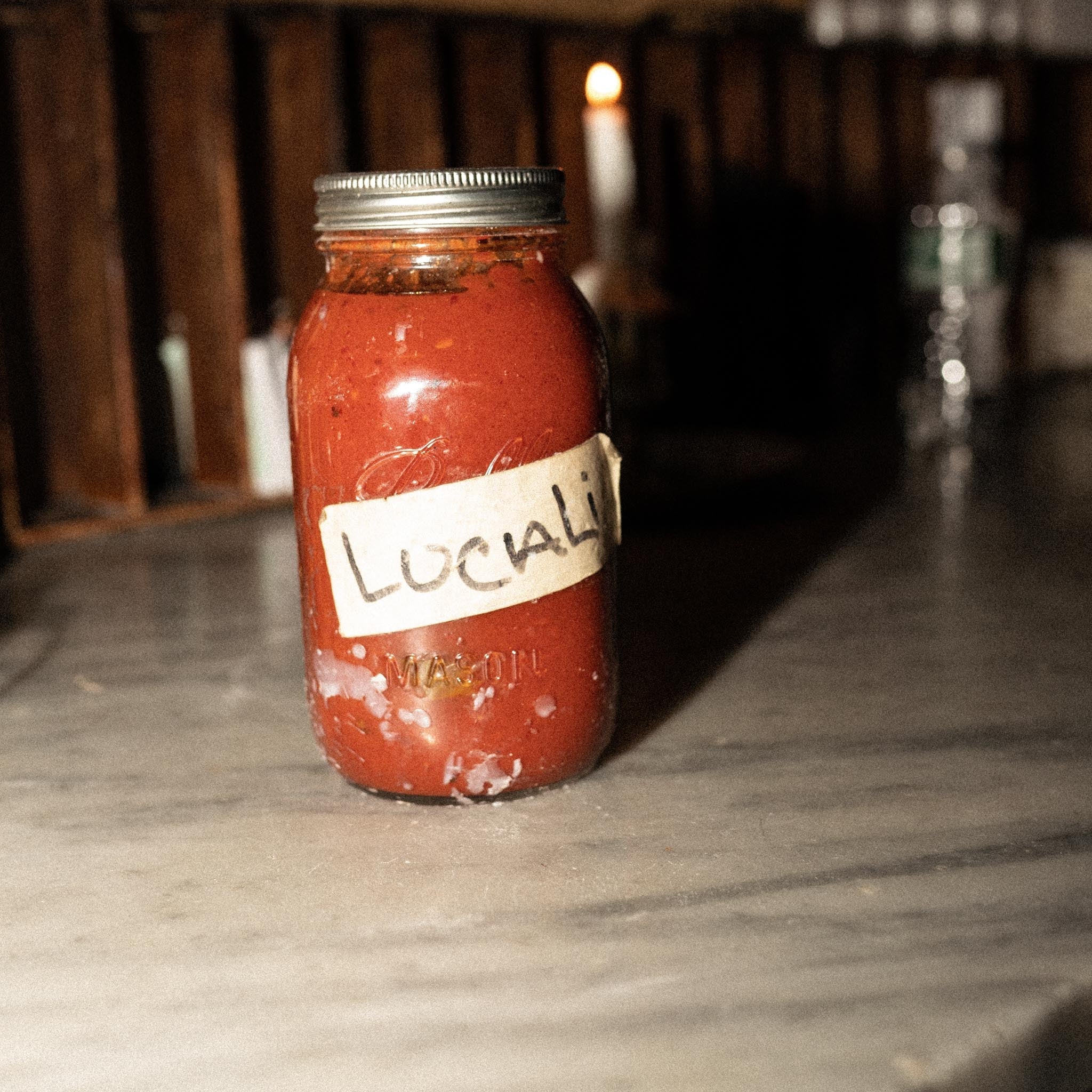 Front view of mason jar of tomato sauce labeled Lucali, showing handwritten label and metal lid on marble counter with candlelit background