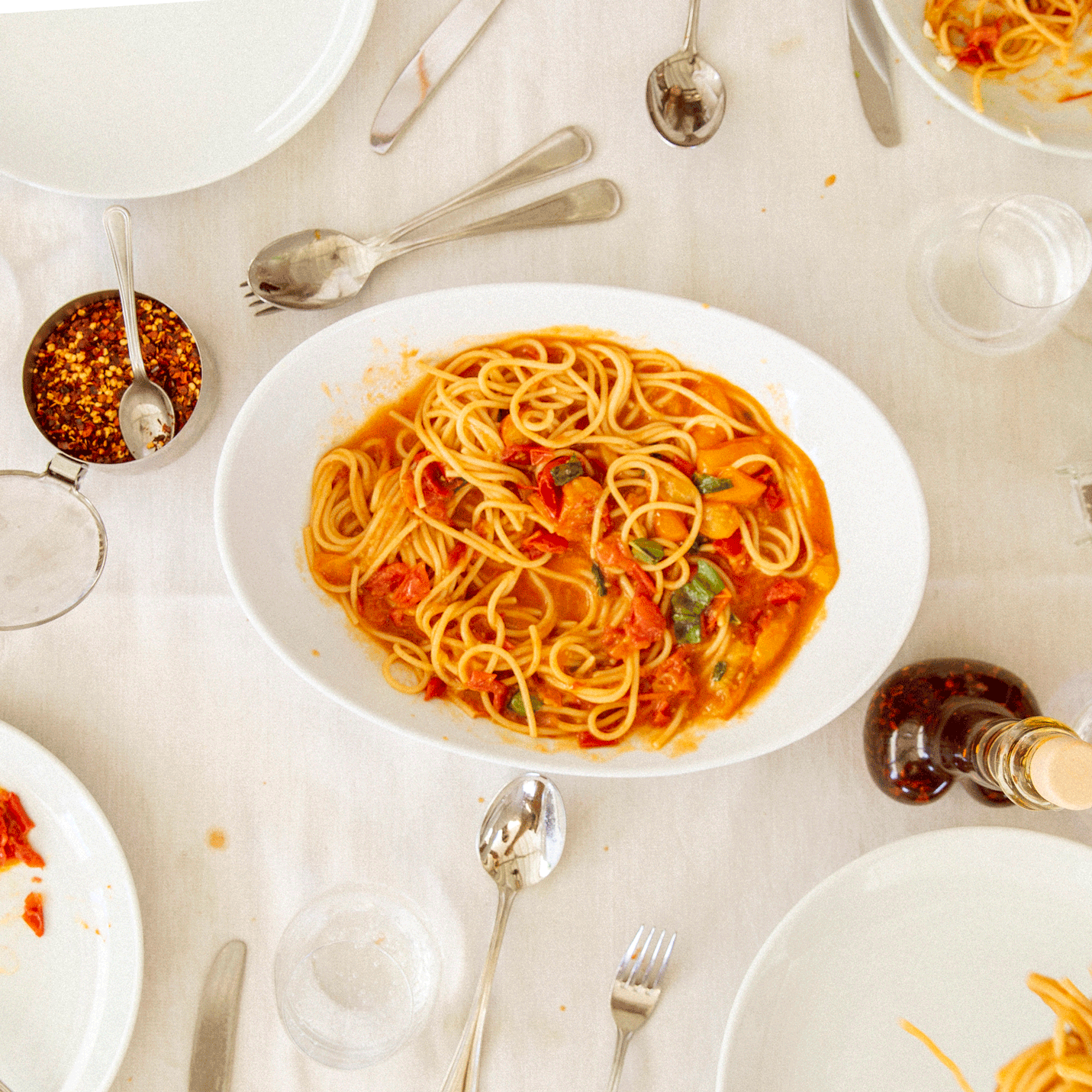 Overhead view of a bowl of spaghetti in tomato sauce with basil on a white linen table surrounded by plates, utensils, and a jar of chili flakes