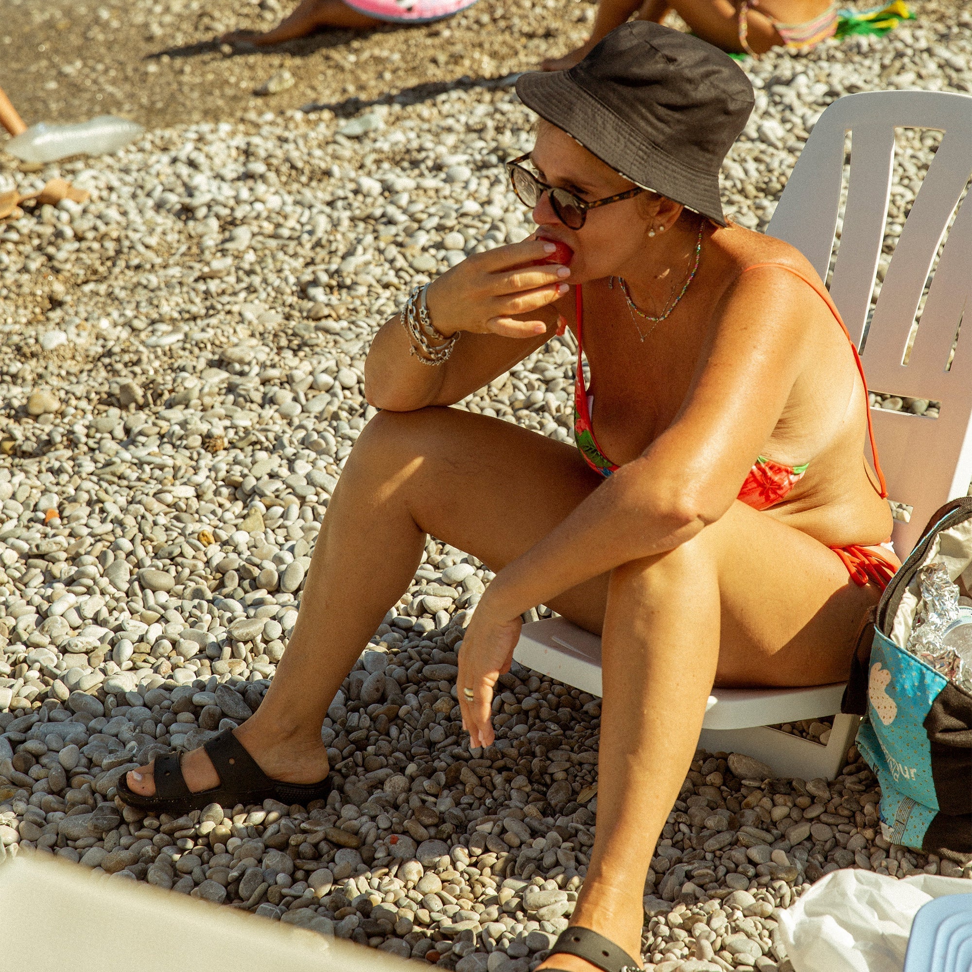 Woman in a floral red bikini and black bucket hat sitting on a plastic beach chair eating a peach on a pebbled beach