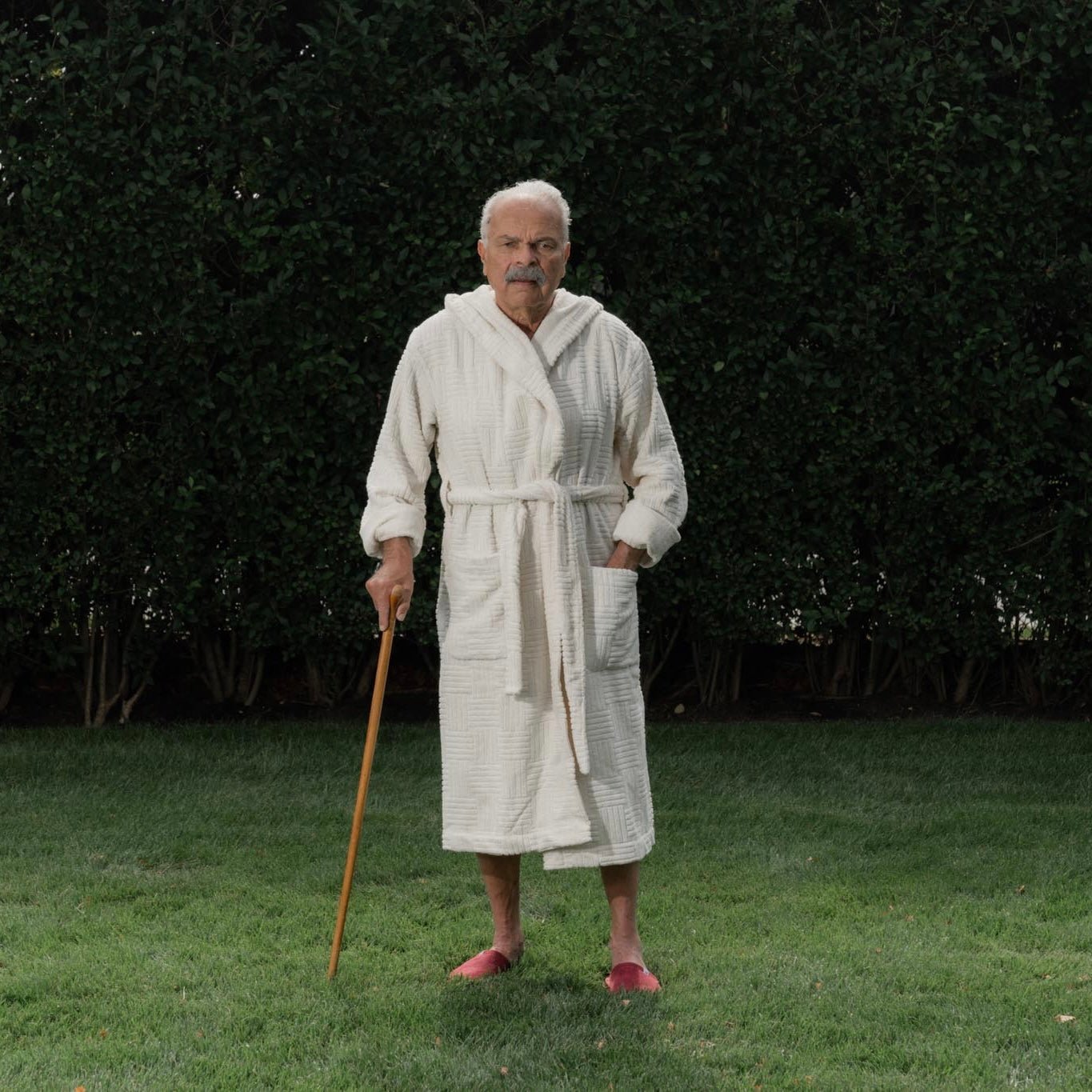 Man standing on grass wearing a white textured robe and red slippers, holding a wooden cane in front of a hedge