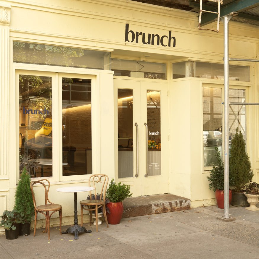 Brunch storefront with cream facade, glass doors, and two sidewalk café chairs and table in Soho, New York