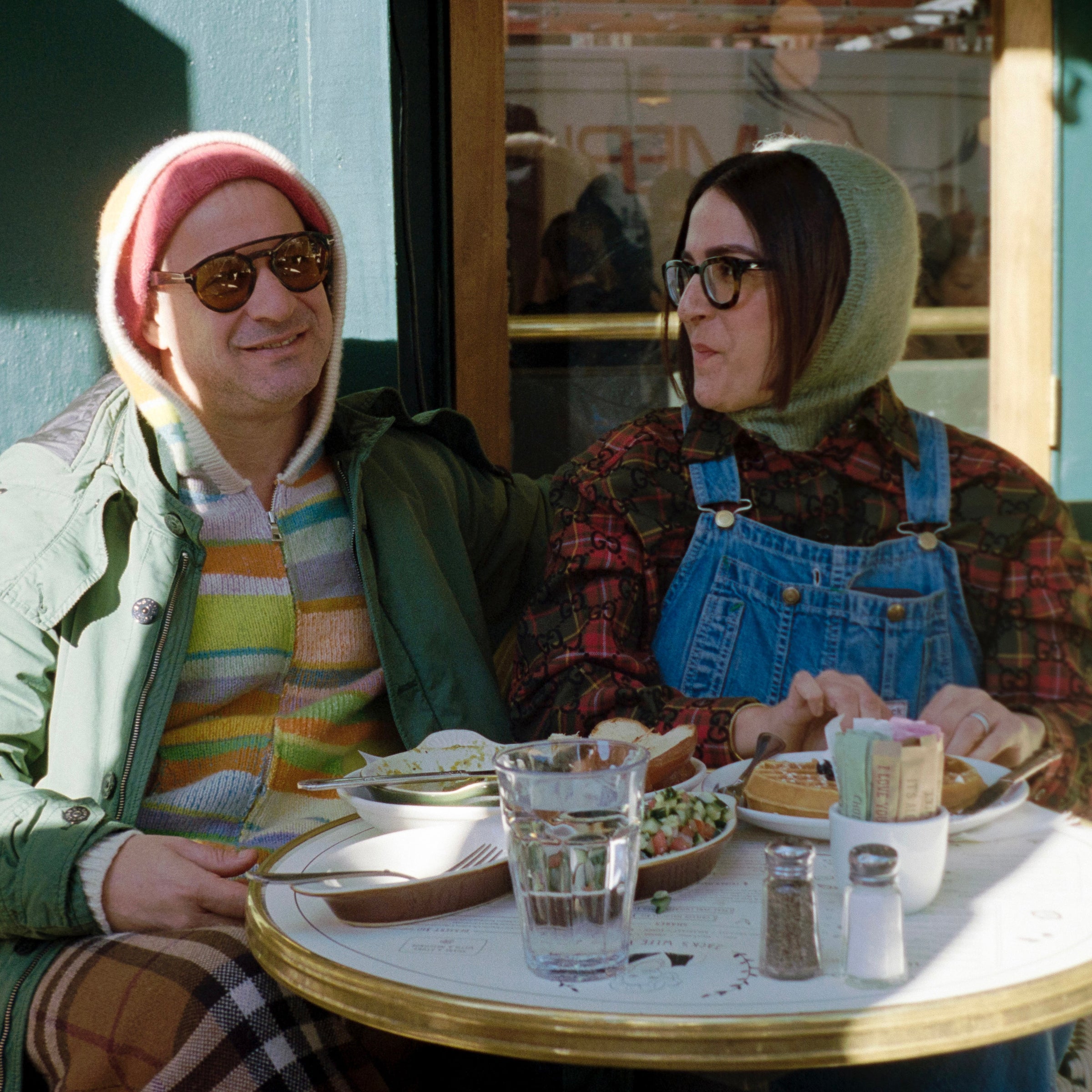Couple at sidewalk café sharing a meal; man in sunglasses and striped hoodie, woman in glasses and denim overalls.