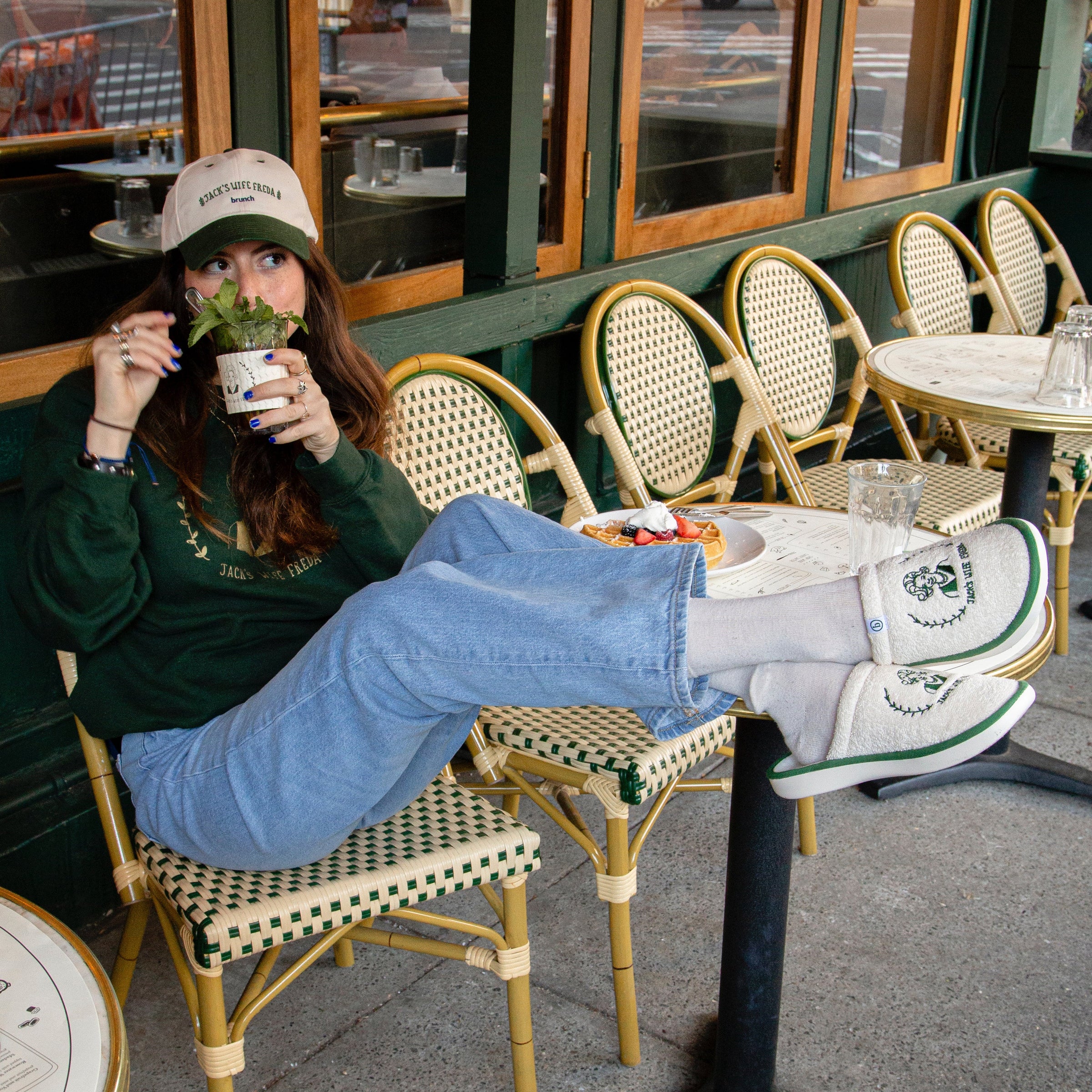 Woman lounging at sidewalk café wearing cap and sweatshirt reading 'JACK'S WIFE FREDA brunch'.