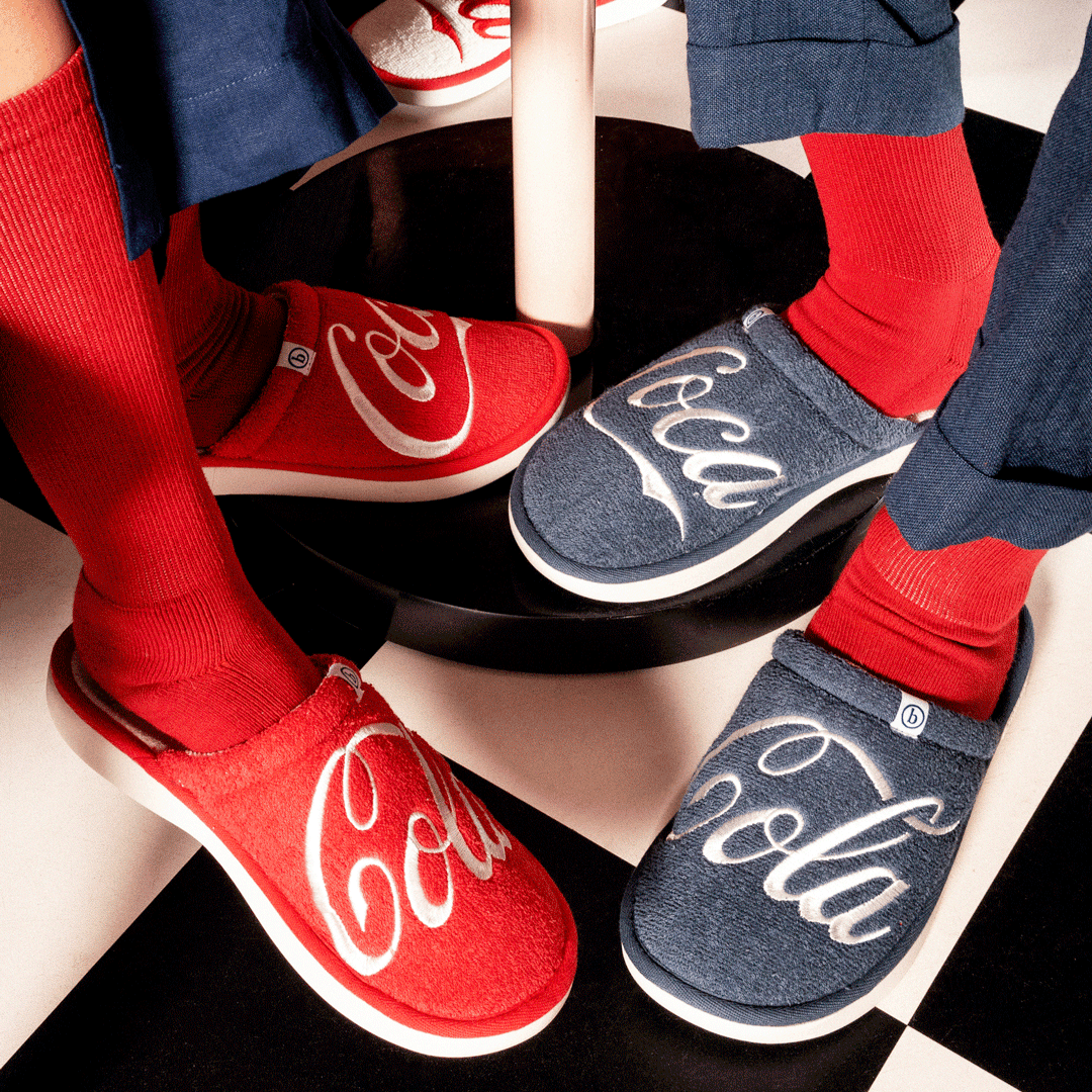 Top-down view of Brunch x Coca-Cola red and navy terry slippers on socks, white Coca-Cola script on checkered floor