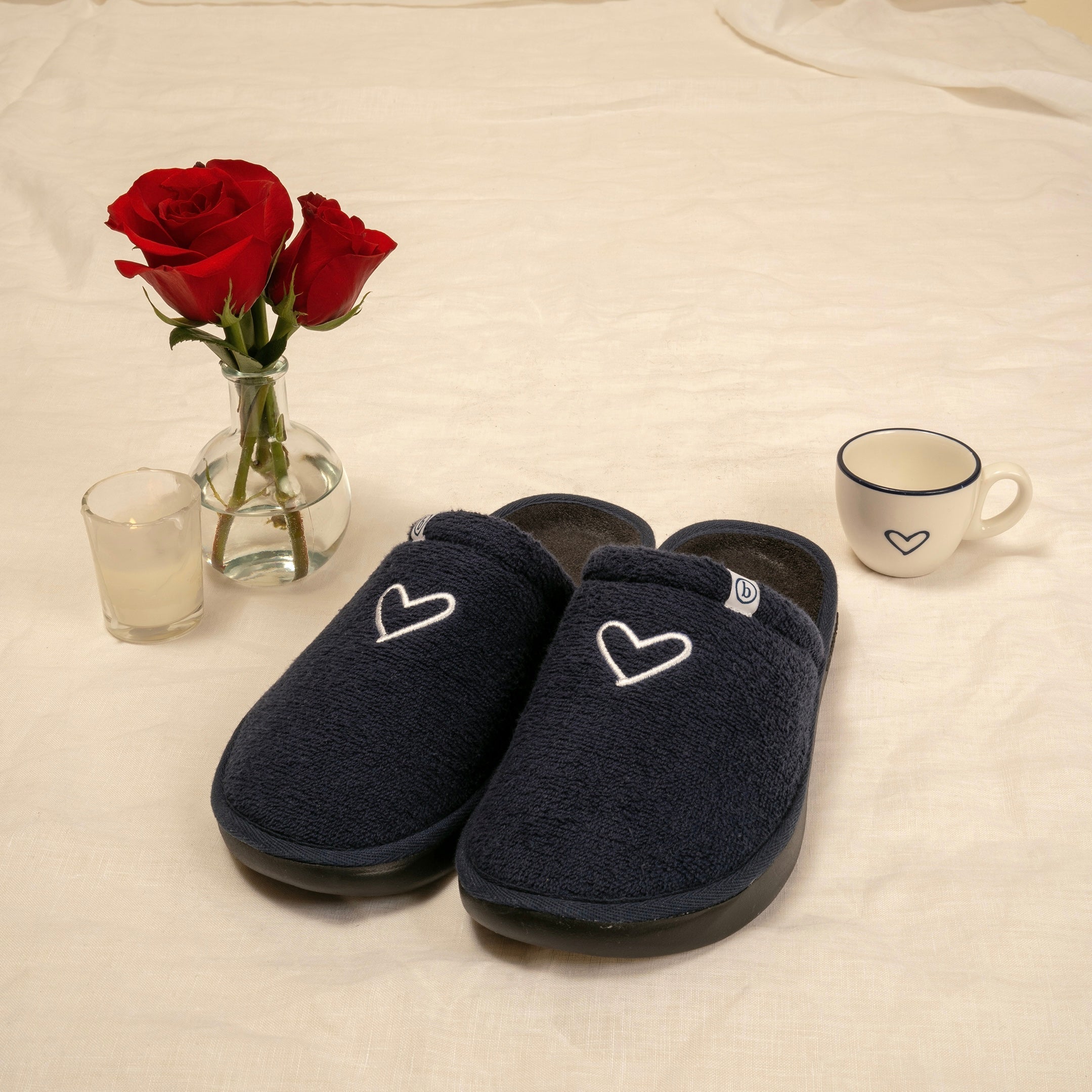 Front three-quarter view of navy Brunch l’Essentiel slippers showcasing embroidered white heart, contoured footbed, and thick outsole on cream linen with roses and cup