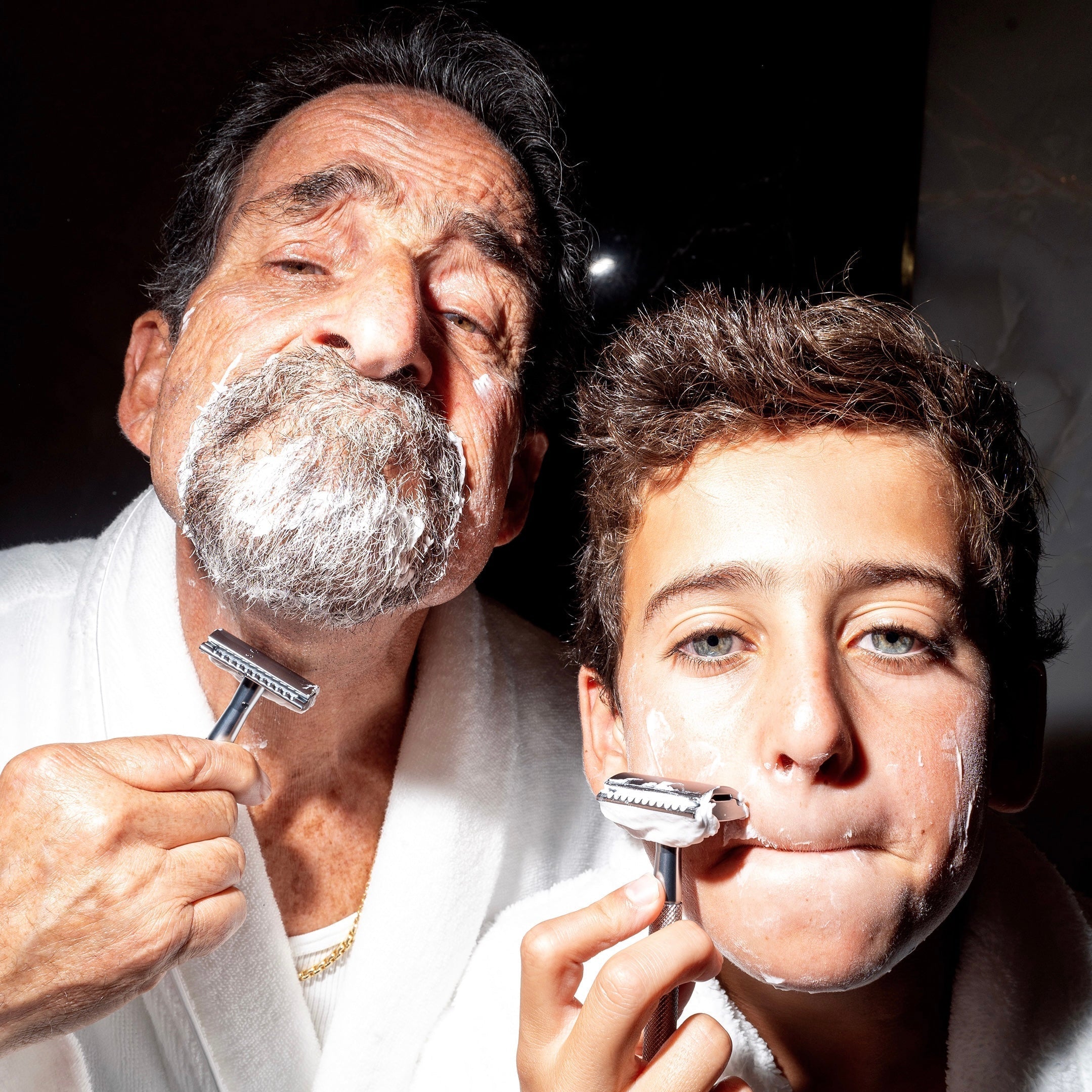 Older man and young boy pretending to shave together in white robes, both with shaving cream and safety razors, close-up