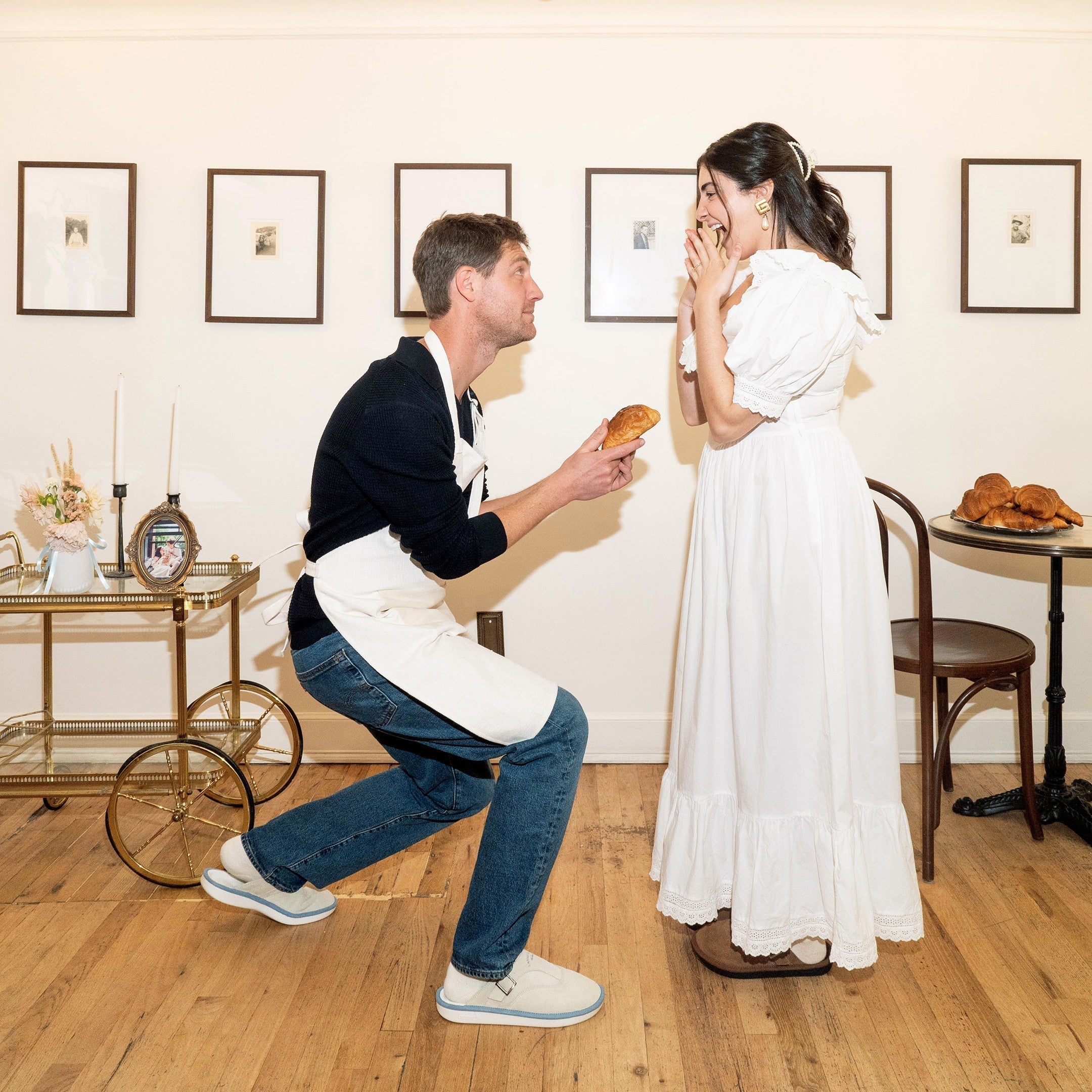 Man in apron kneeling to offer a croissant to a surprised woman in a white dress in a gallery room