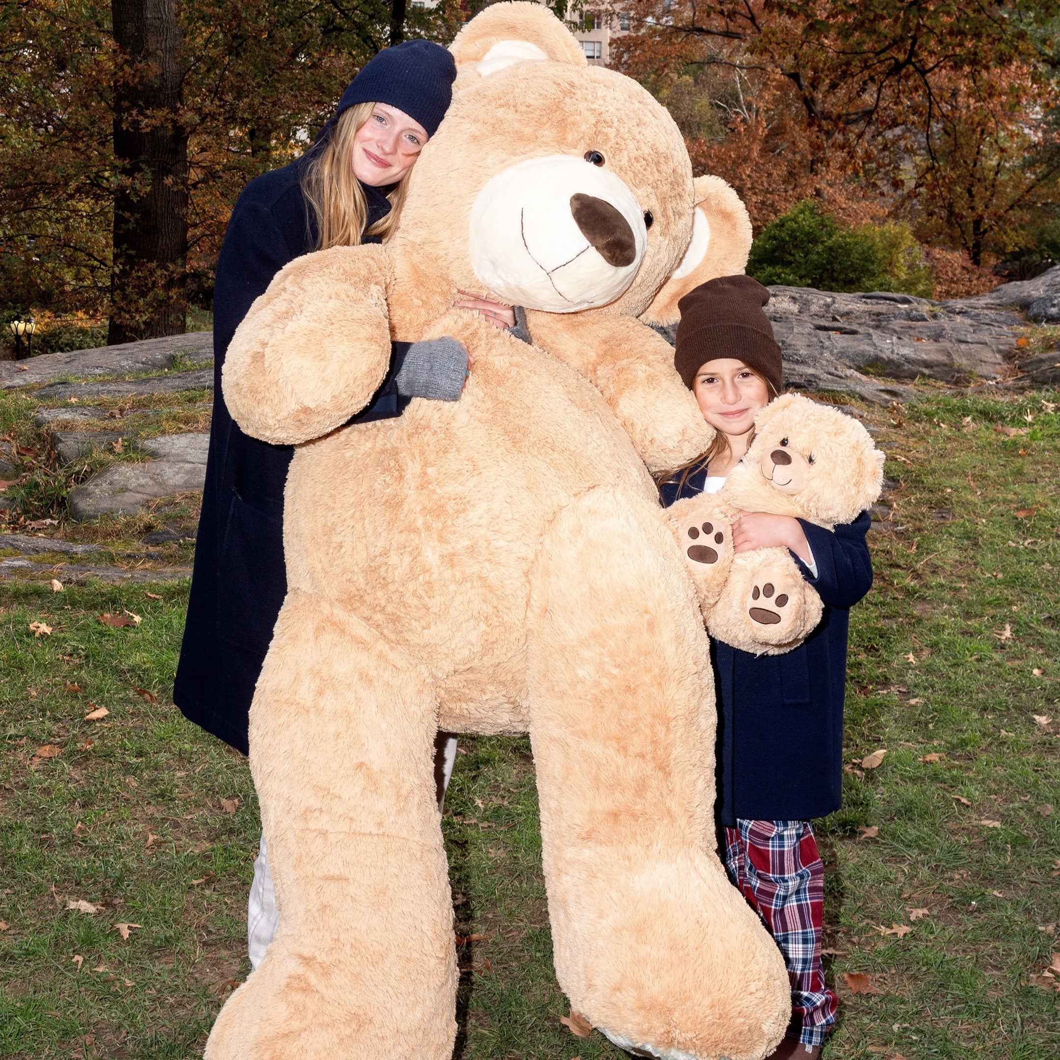 Adult and child wearing beanies in an autumn park hugging a giant teddy bear; child holds a smaller teddy.