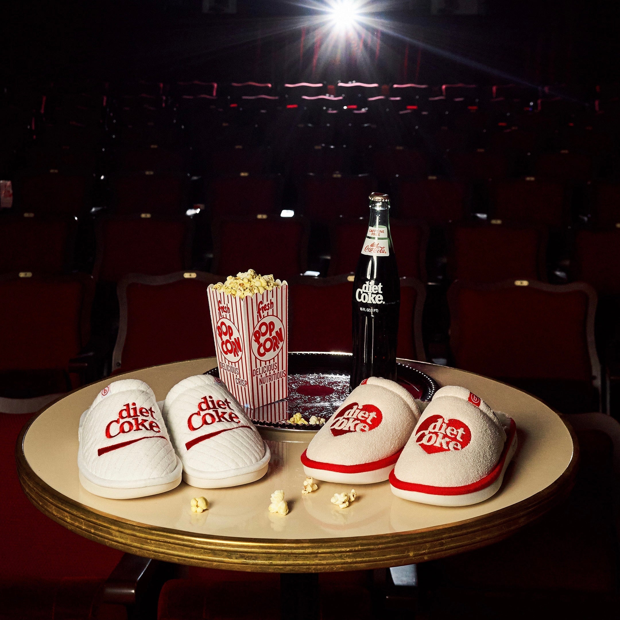 Movie-theater table with striped popcorn box reading 'fresh POP CORN', a bottle labeled 'diet Coke', and two 'diet Coke' slippers.