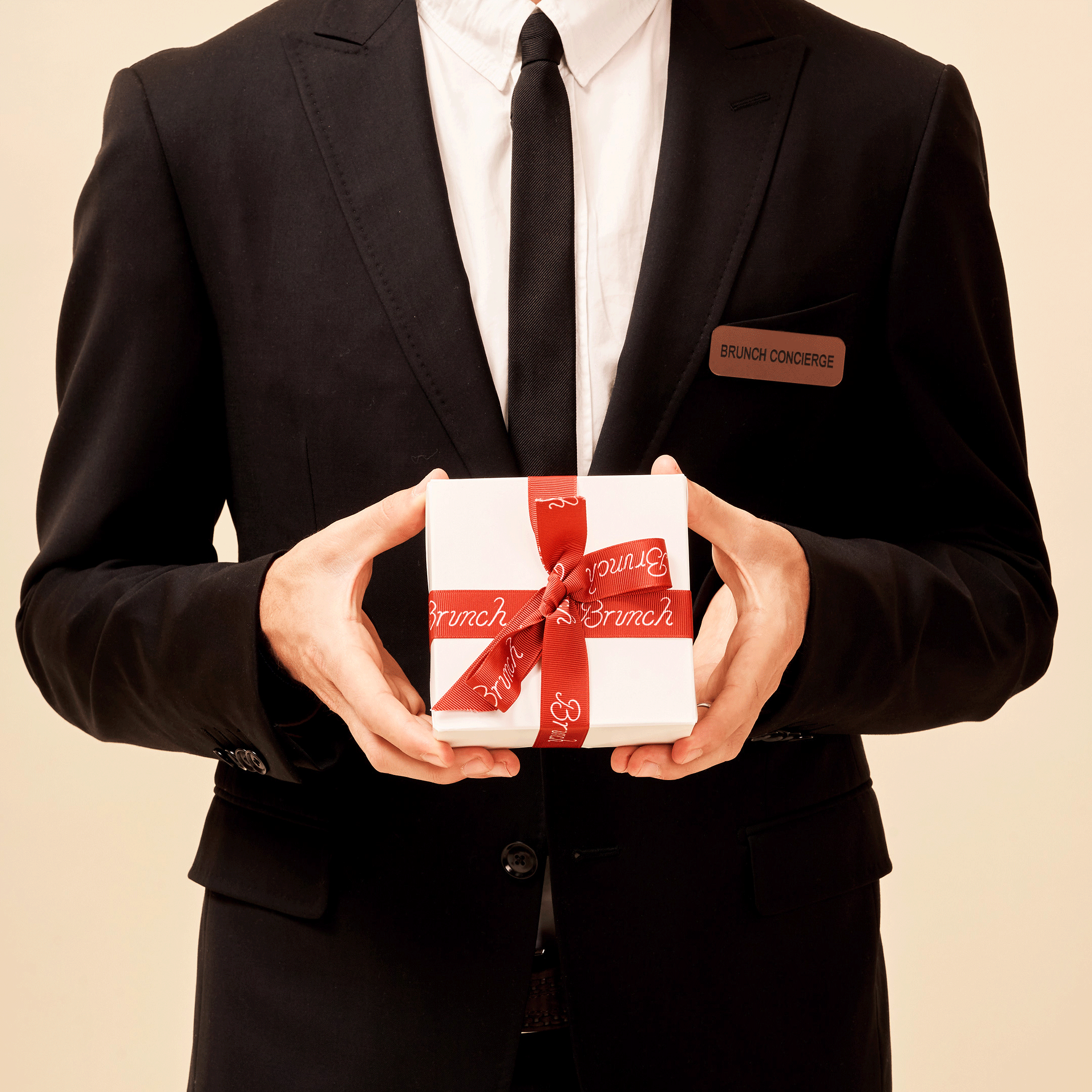 Man in black suit holding a white gift box tied with a red ribbon printed Brunch, wearing a name badge reading Brunch Concierge
