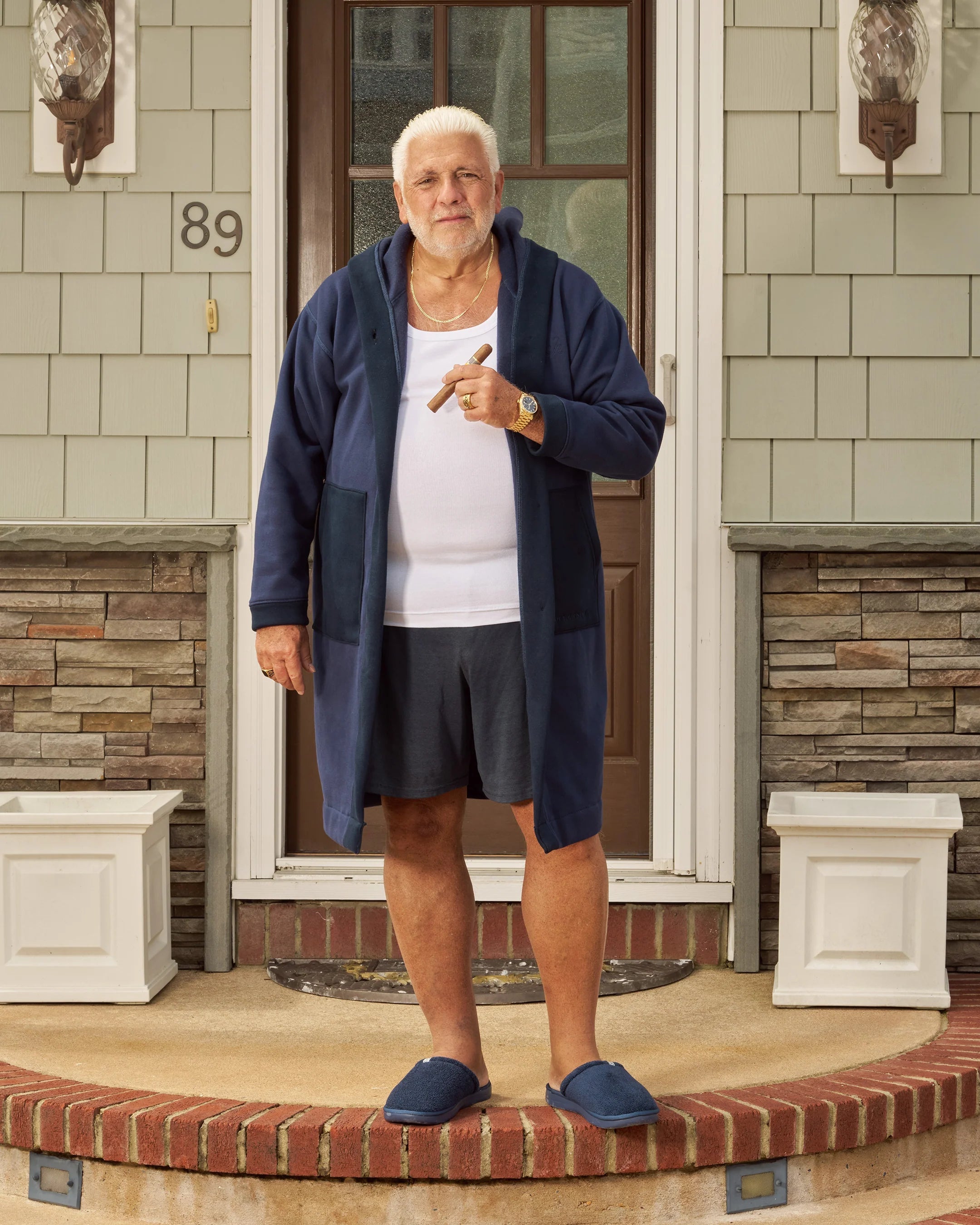 Older man on front porch (house number 89) wearing navy robe coat, white tank, holding a cigar