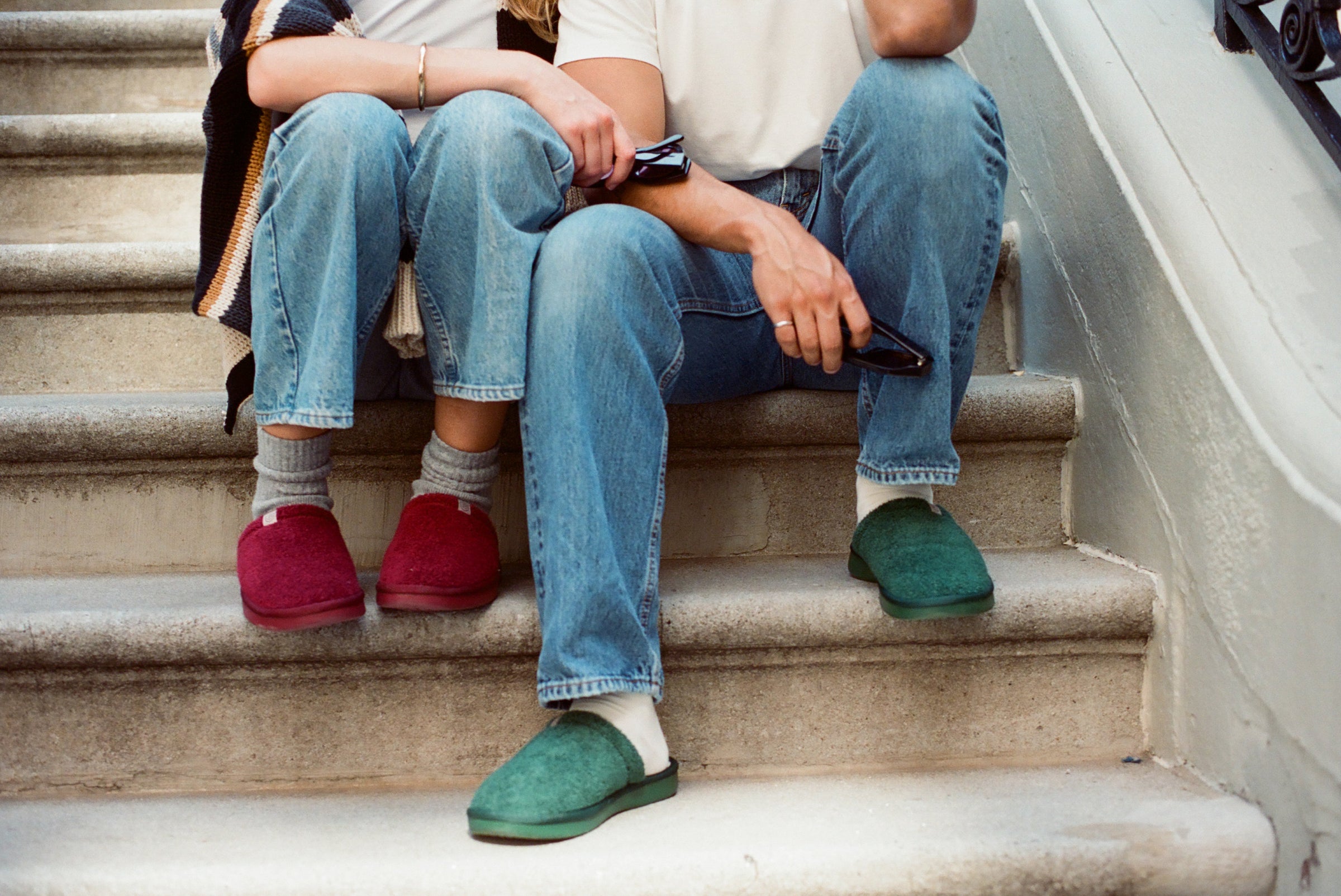 Two people sitting on a stoop wearing jeans, one with red felt slip-on slippers and the other with green slip-on slippers