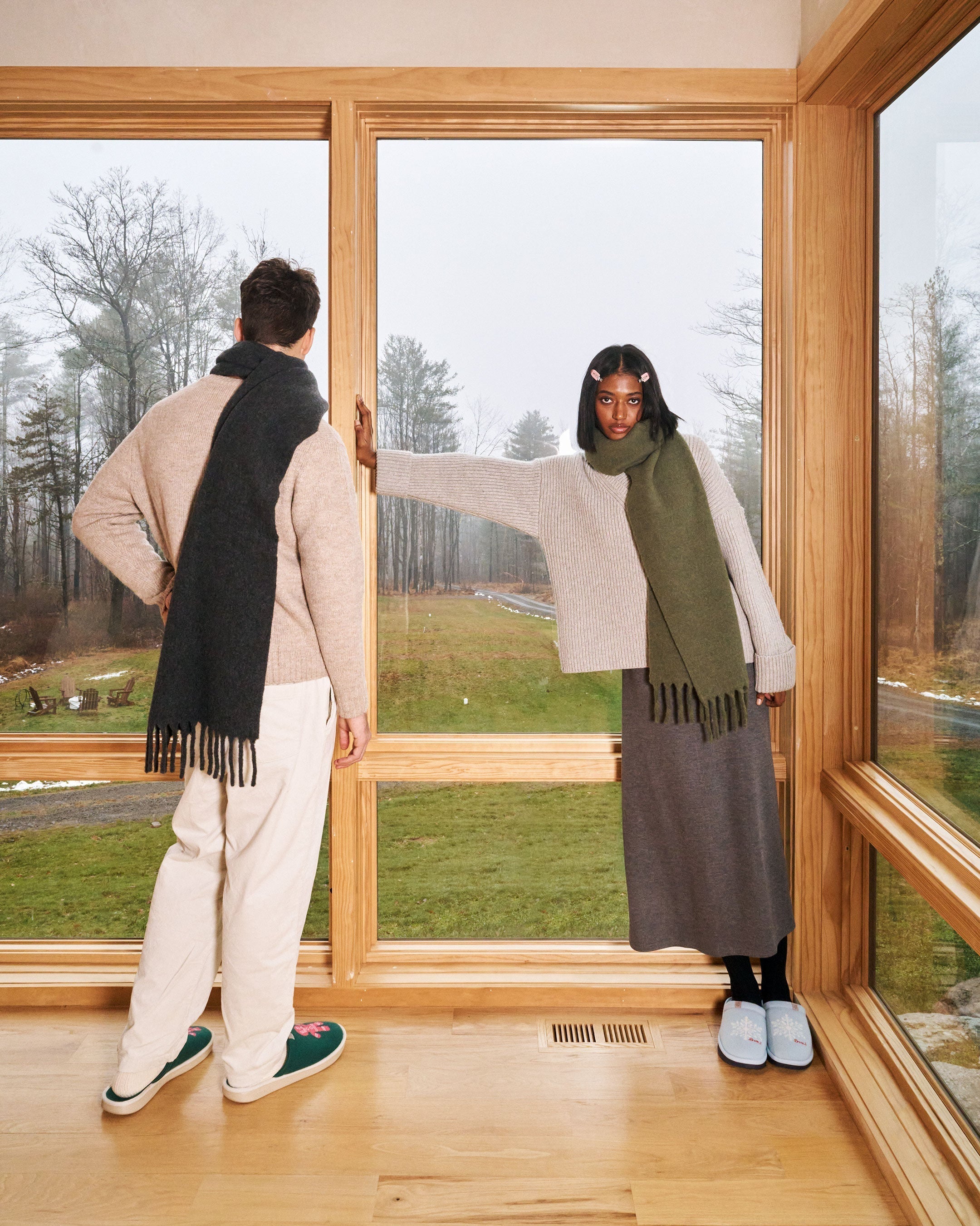 Two models wearing Essential Cashmere slippers and knitwear in a wood cabin by a large window overlooking snowy trees