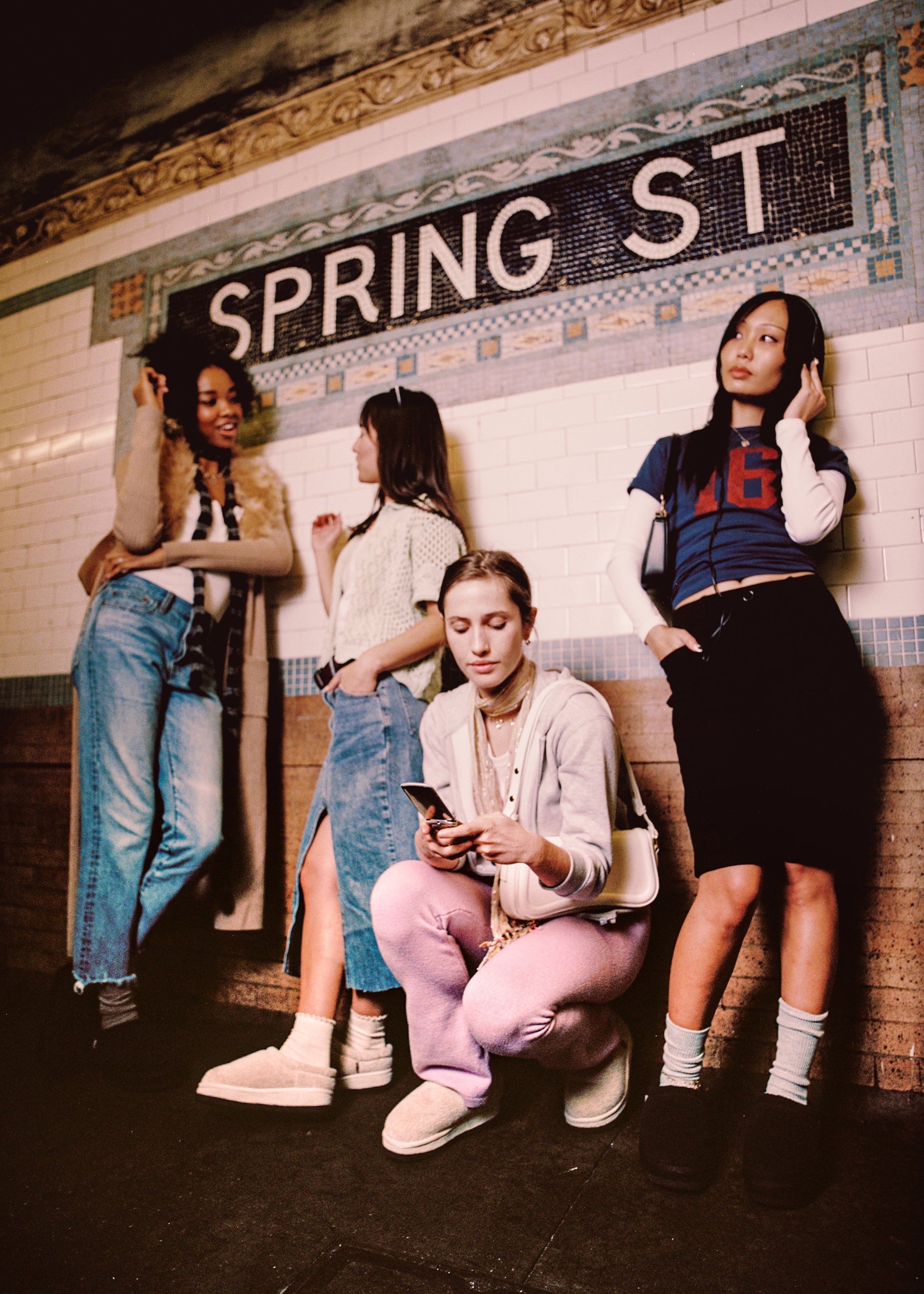Four young women at tiled SPRING ST subway mosaic; one crouches using a phone, another wears a blue shirt reading 16.