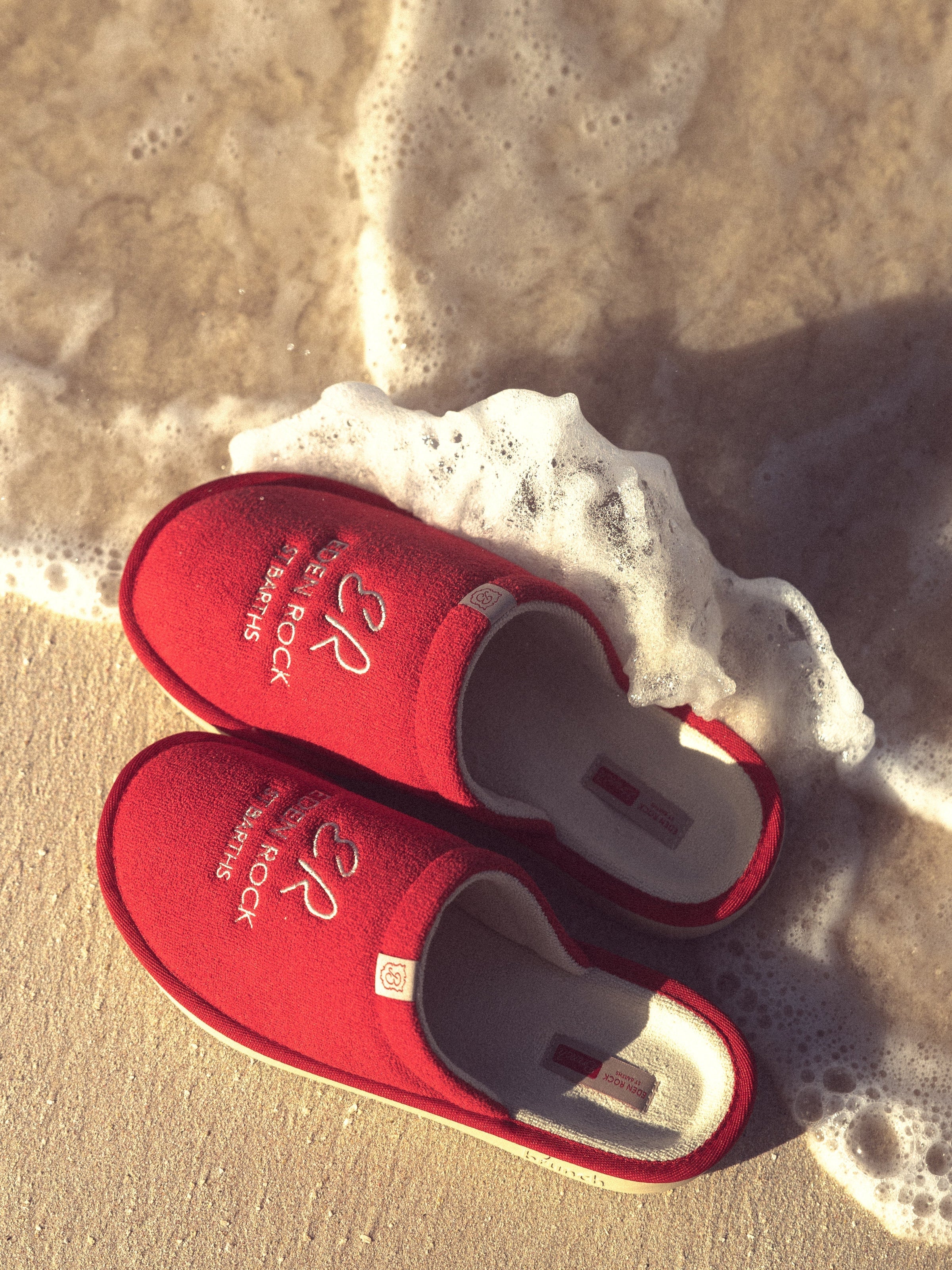 Top view of red Eden Rock St Barths slippers with embroidered logo and cushioned interior on sandy beach with sea foam