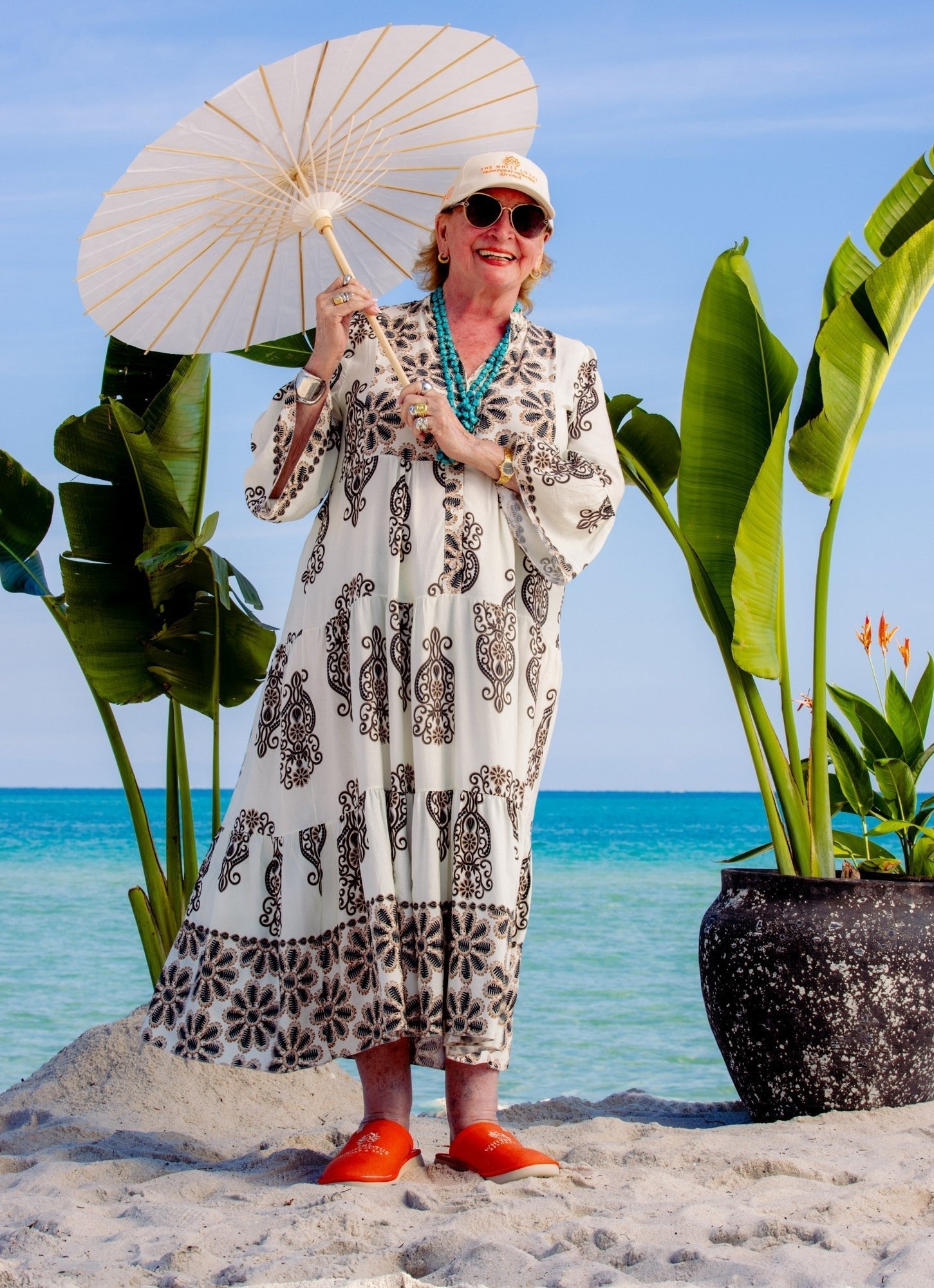 Older woman on a sandy beach holding a white parasol, wearing a printed dress, turquoise beads, and orange slippers