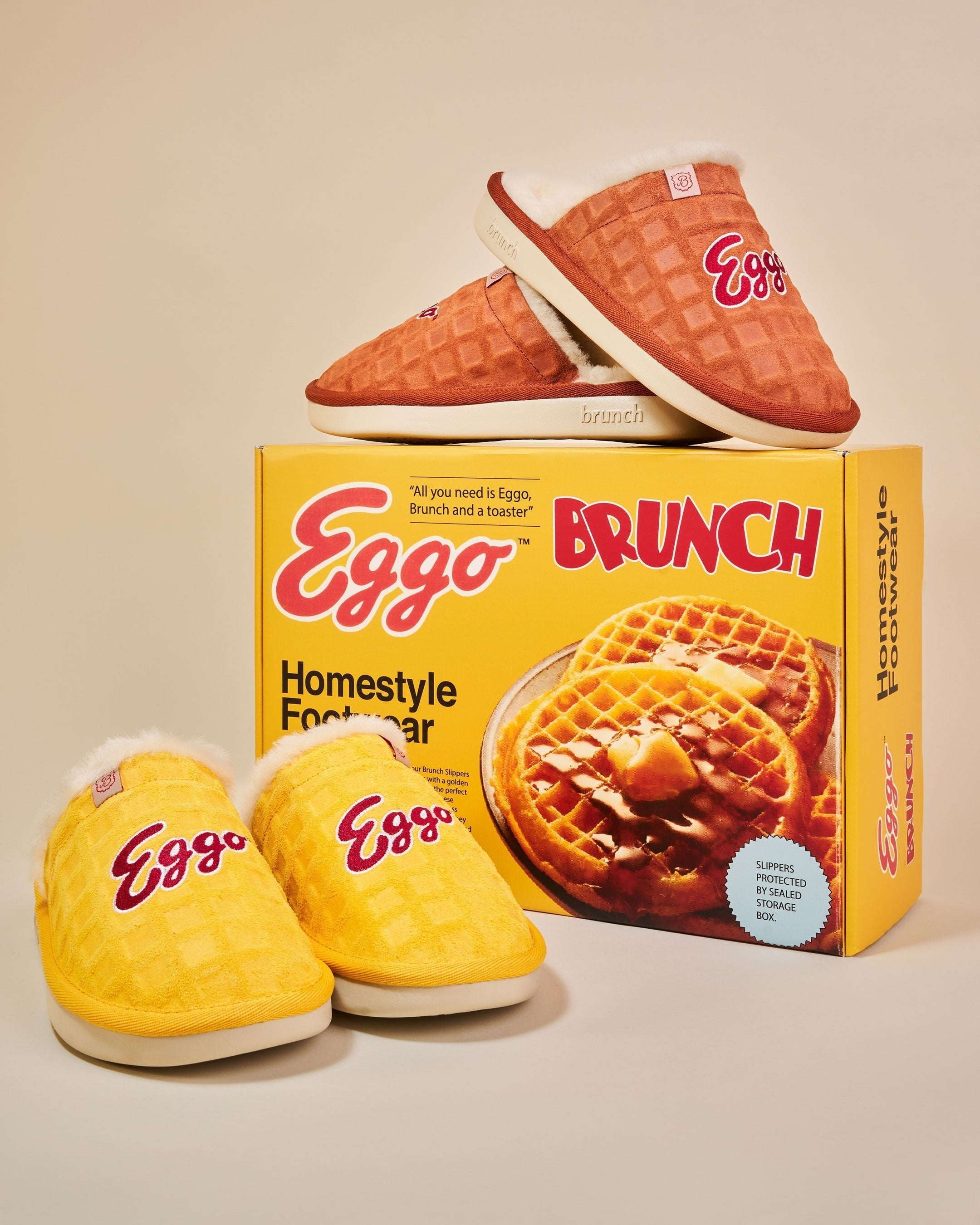Three-quarter view of Eggo slippers with waffle-textured upper and embroidered Eggo logo, yellow Eggo box behind
