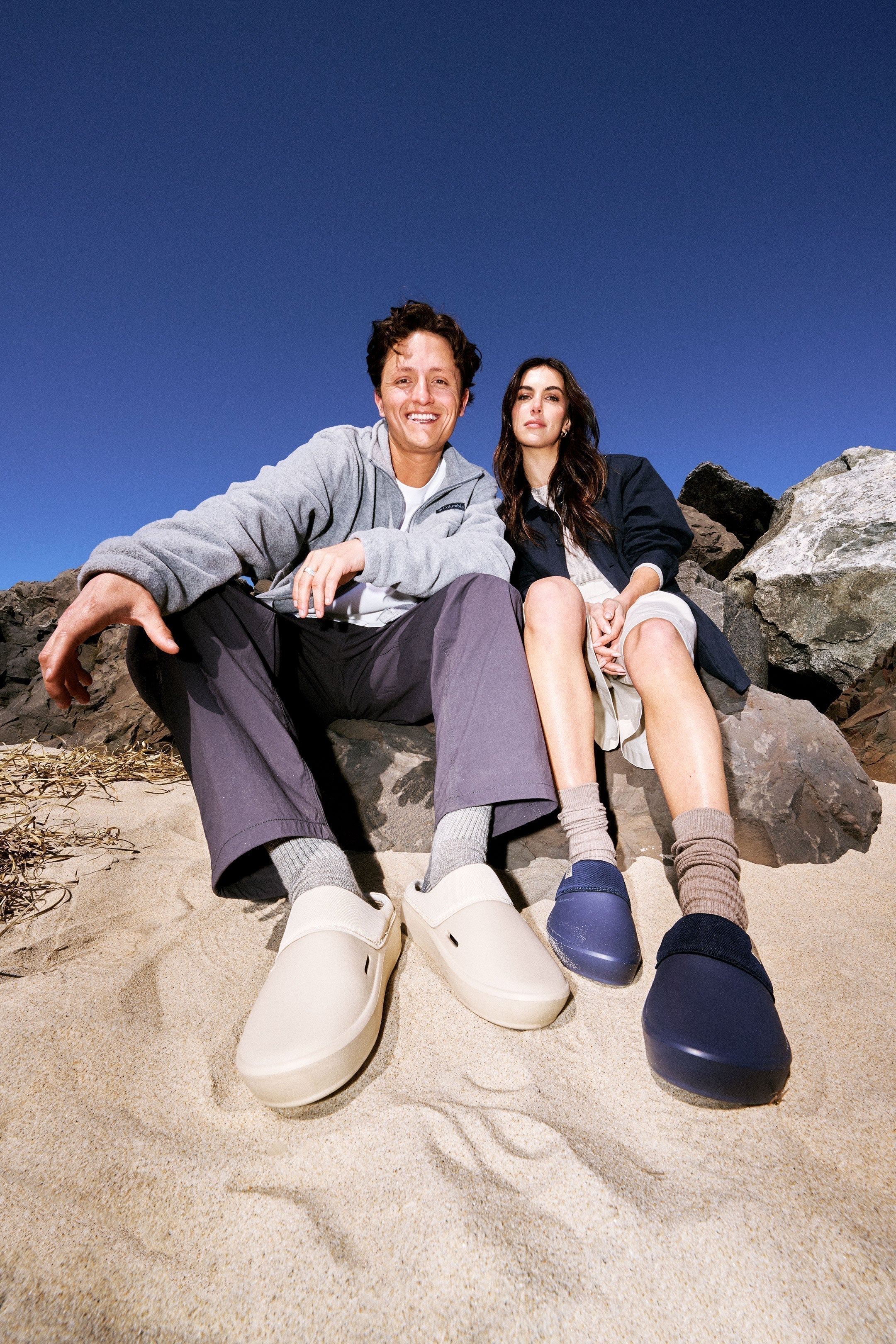 Two people sitting on sand and rocks wearing Aura Clogs, beige pair left and navy pair right beneath a clear blue sky