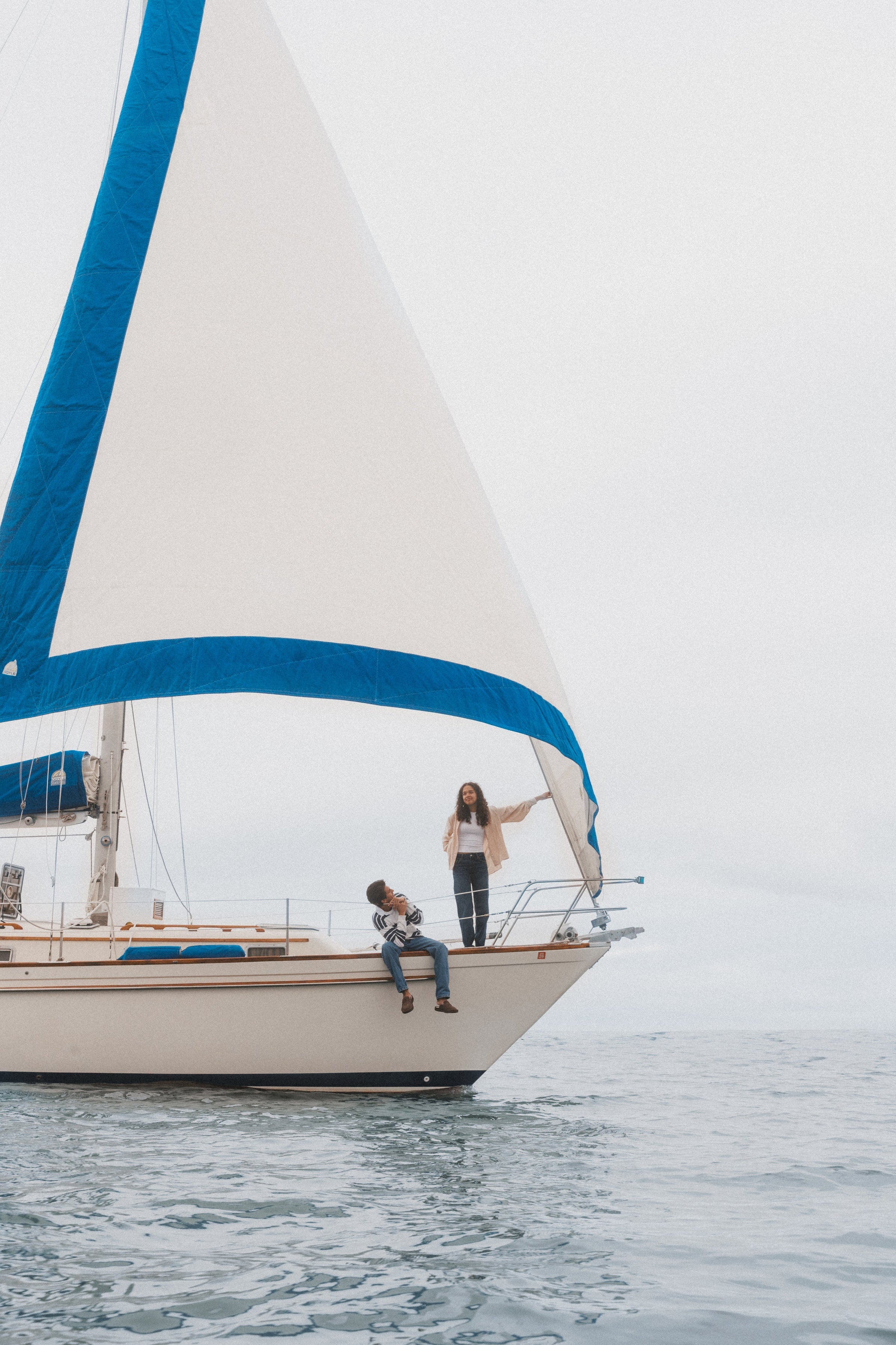 Two people on the bow of a sailboat under a large white sail with blue trim on a calm, overcast sea