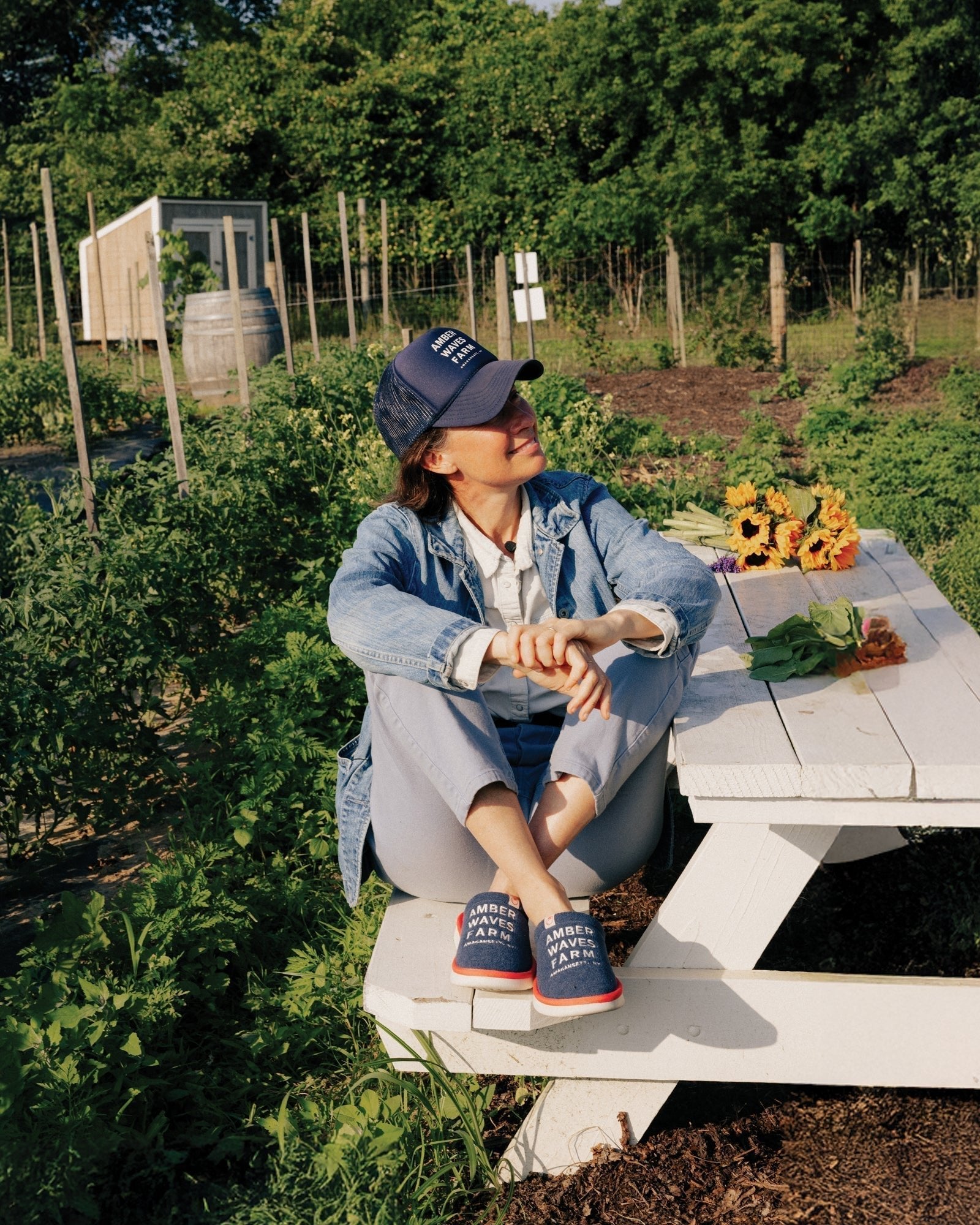Woman seated on a white picnic bench at a farm, wearing a denim jacket, Amber Waves Farm cap and embroidered slippers