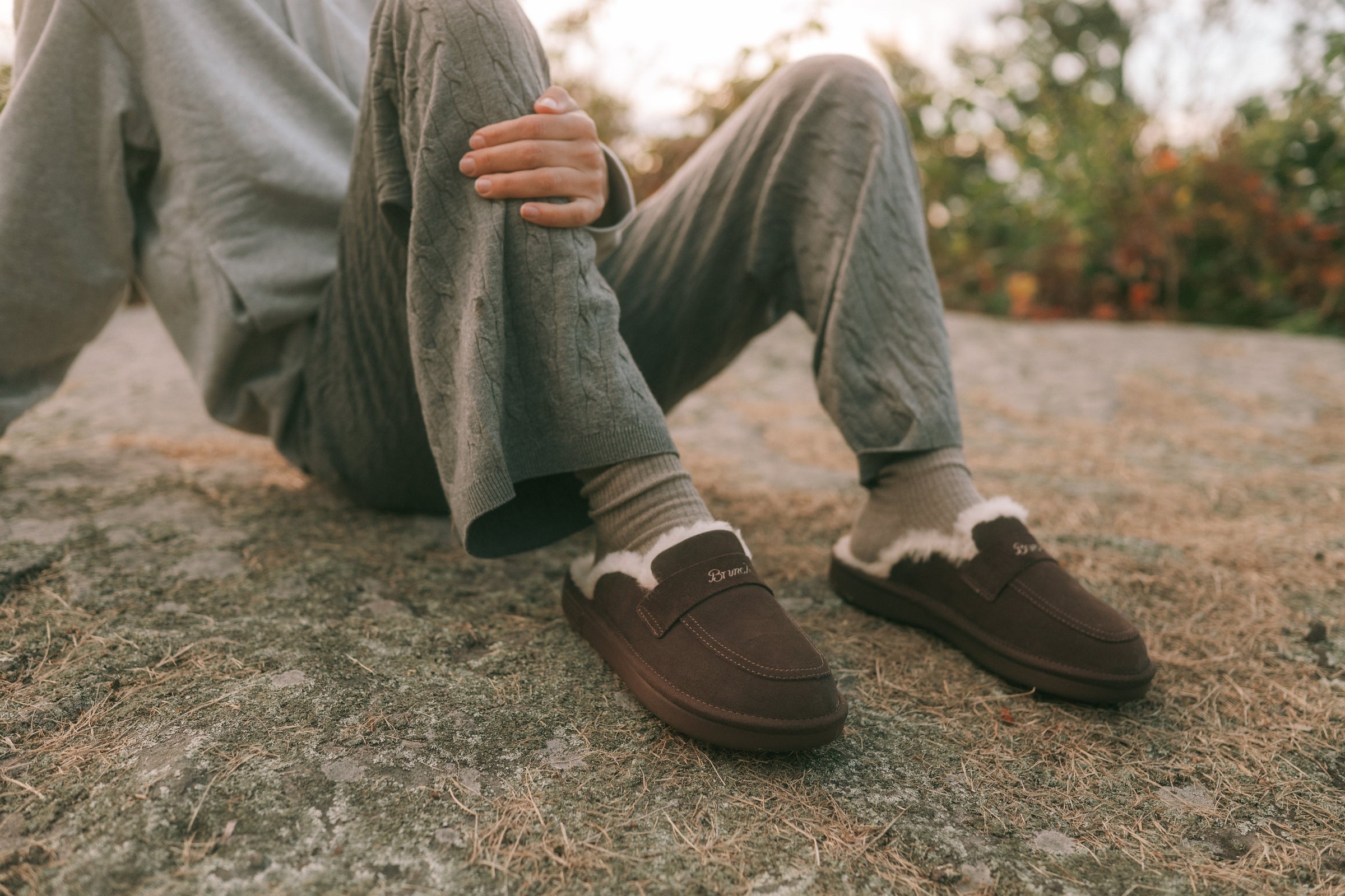 Person seated on a rock wearing brown shearling-lined slip-on loafers and gray knit pants
