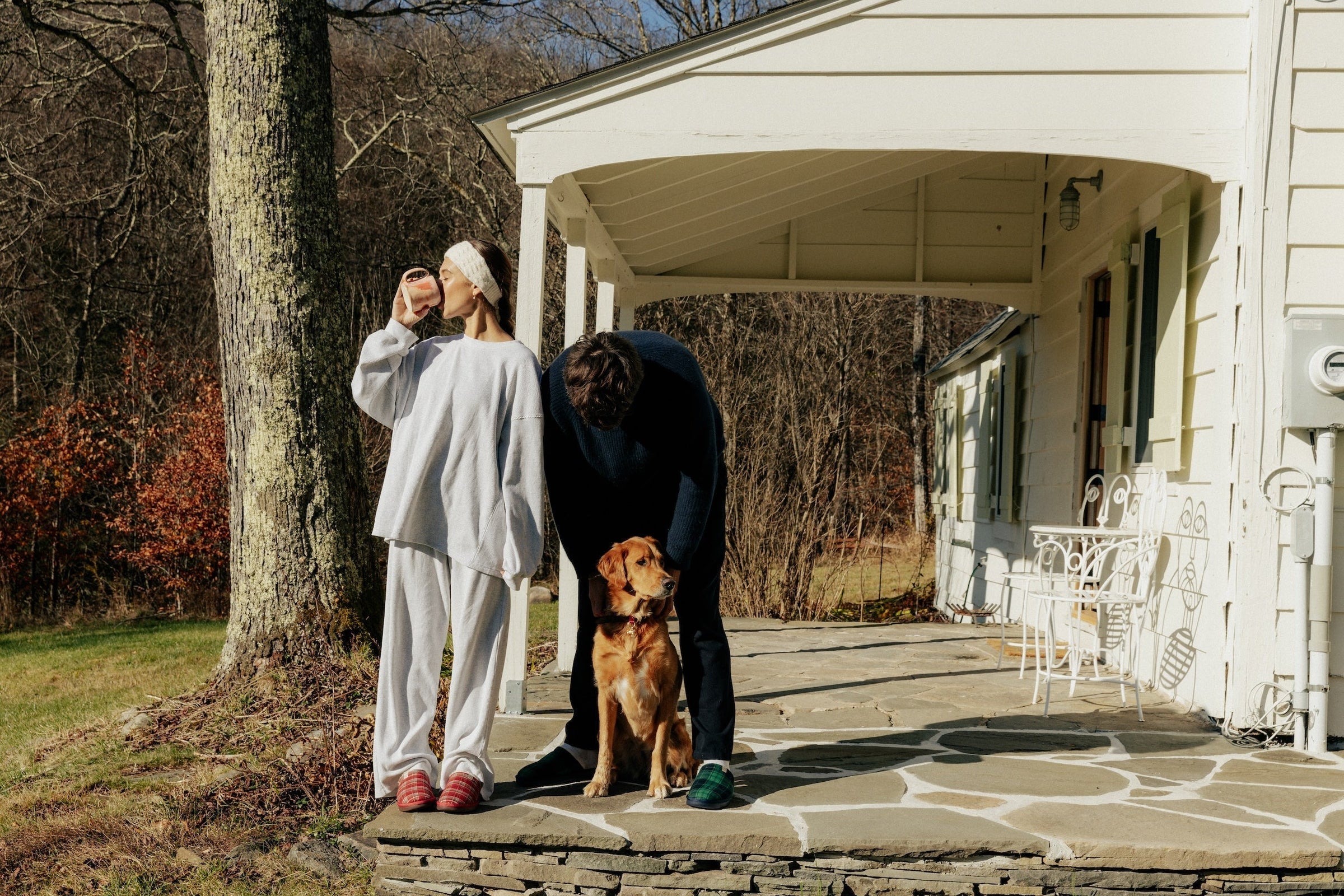 Couple on a cottage porch in cozy loungewear and plaid slippers, man bending to pet a golden retriever