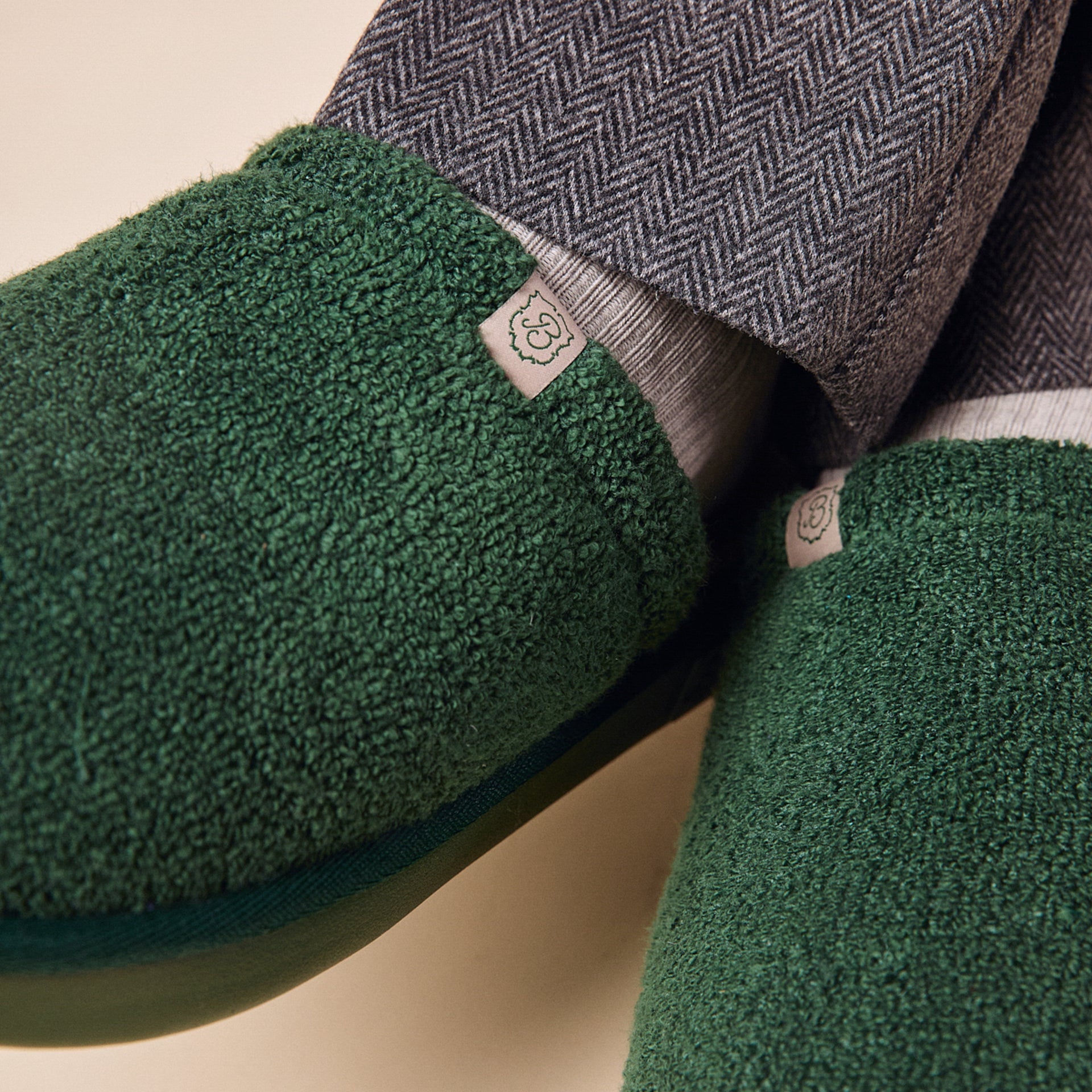 Green sherpa fleece slippers with small fabric logo tag, worn with grey herringbone trousers and light socks.