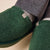 Green sherpa fleece slippers with small fabric logo tag, worn with grey herringbone trousers and light socks.