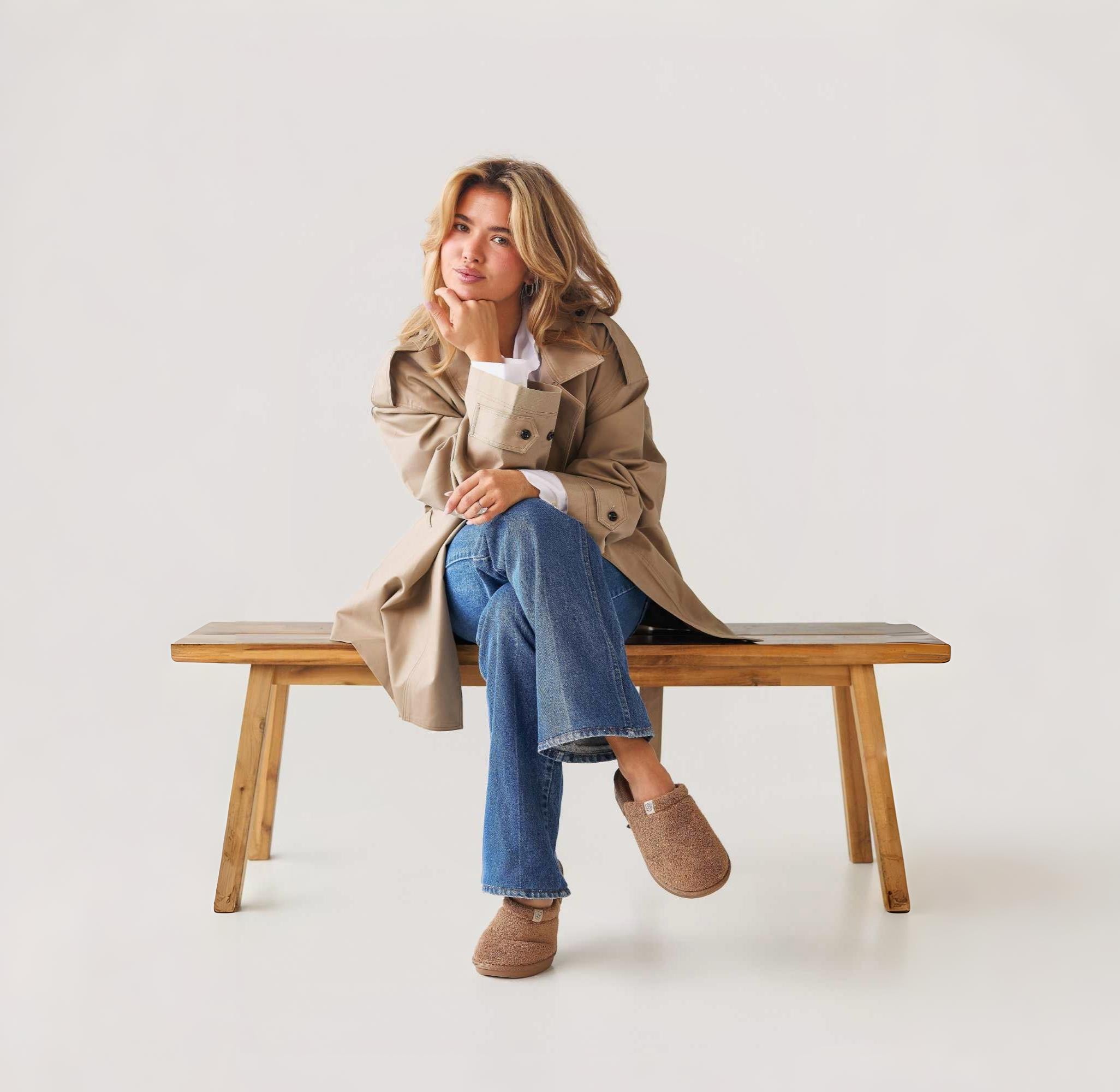 Woman sitting on a wooden bench wearing a trench coat and jeans against a white background