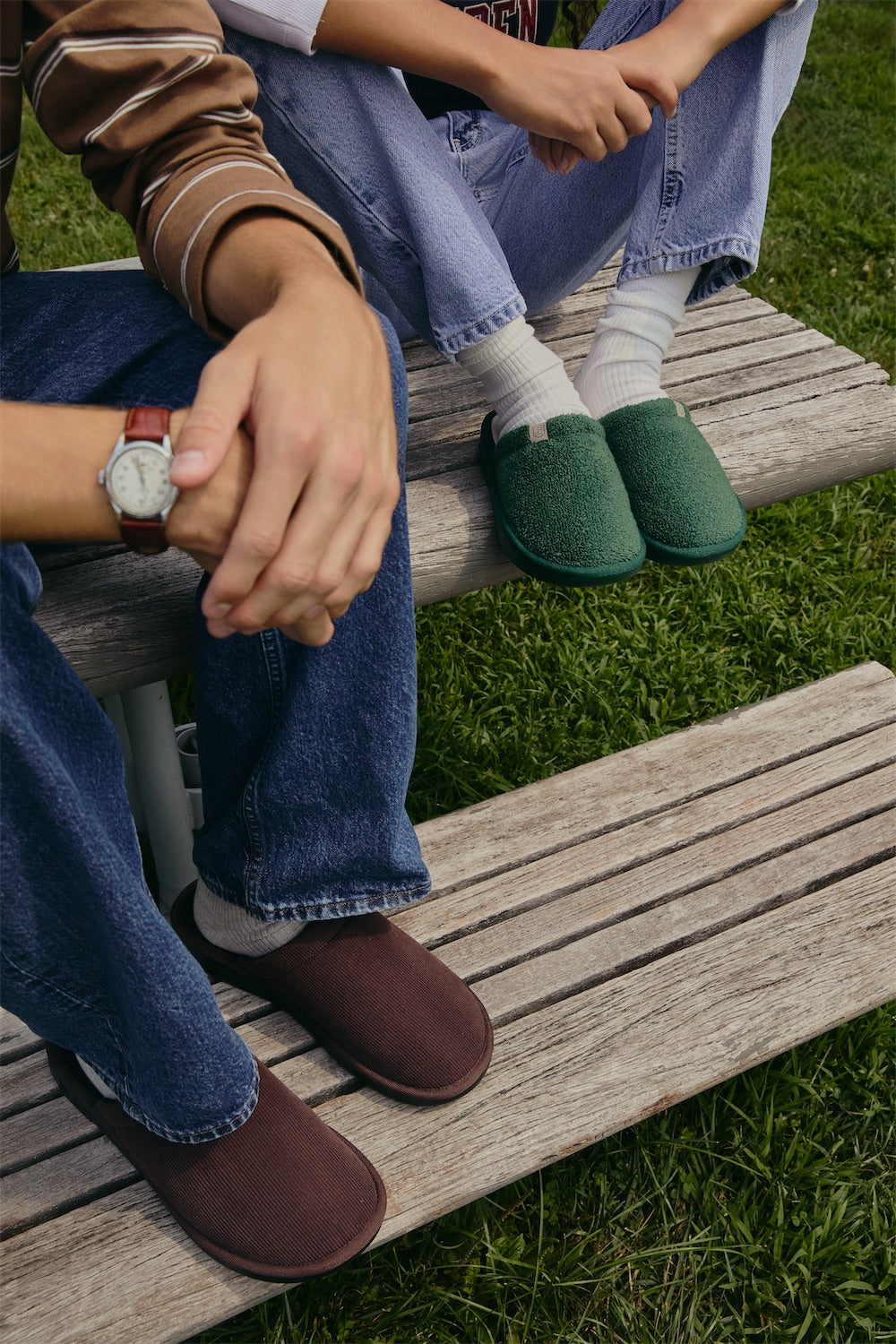 Two people seated on a weathered wooden bench, hands clasped and feet in brown and green slippers over grass.
