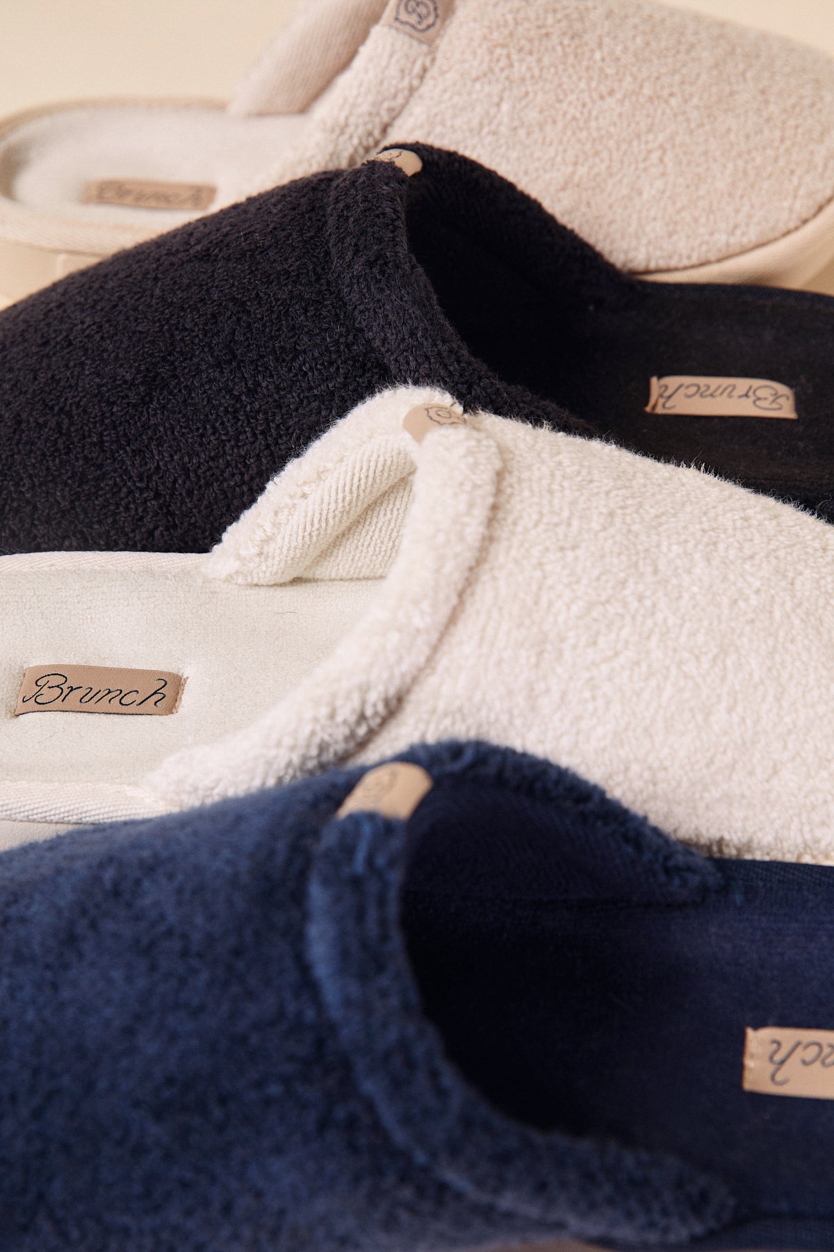 Three terry-cloth slip-on slippers in navy, black, and cream with Brunch leather labels