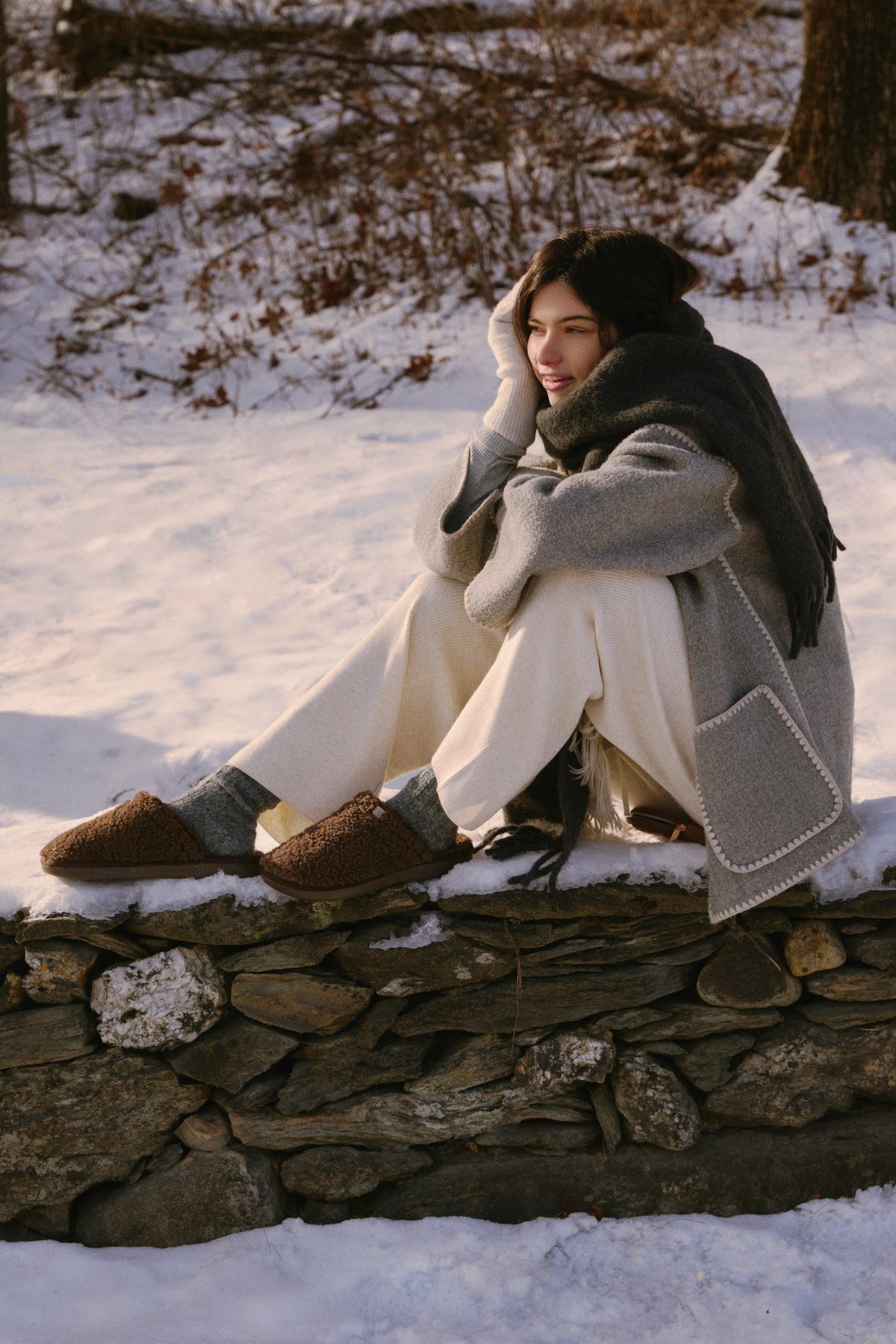 Woman seated on a stone wall in snow, wearing a gray coat and scarf, cream pants and brown slippers.