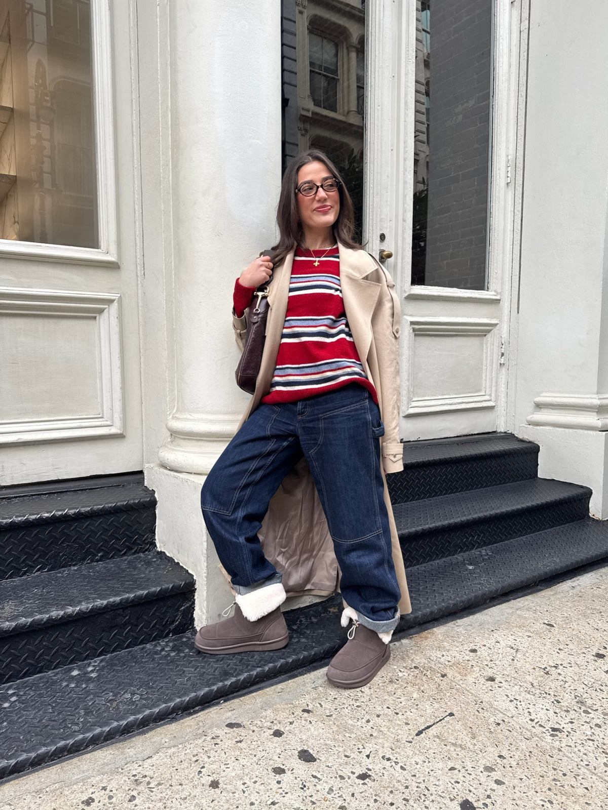 Woman leaning on white stoop wearing beige trench, red striped sweater, cuffed jeans, shearling ankle boots, glasses, smiling.