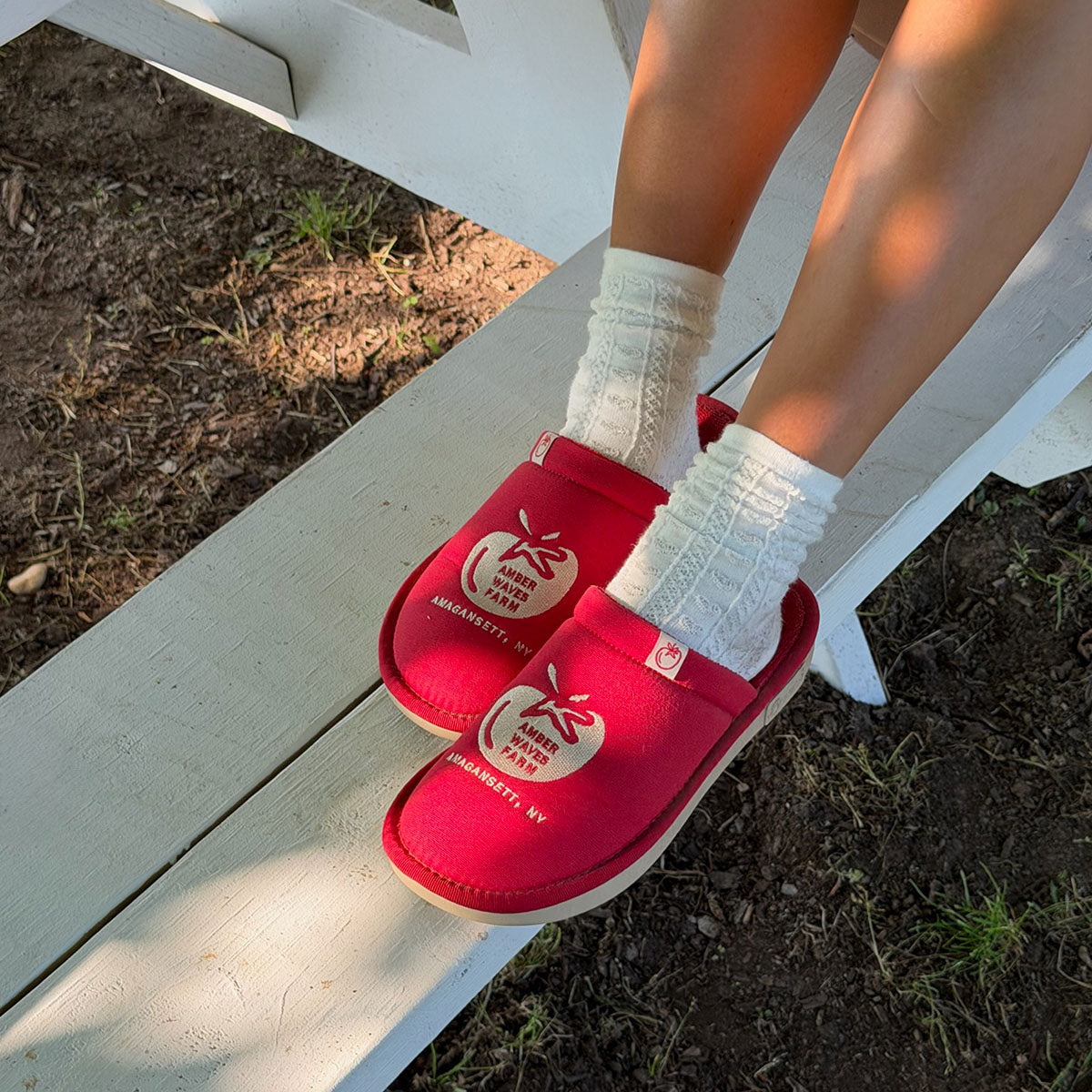 Three-quarter view of Tomato Red Essential slipper showing Amber Waves Farm logo, worn with white socks on a white bench outdoors