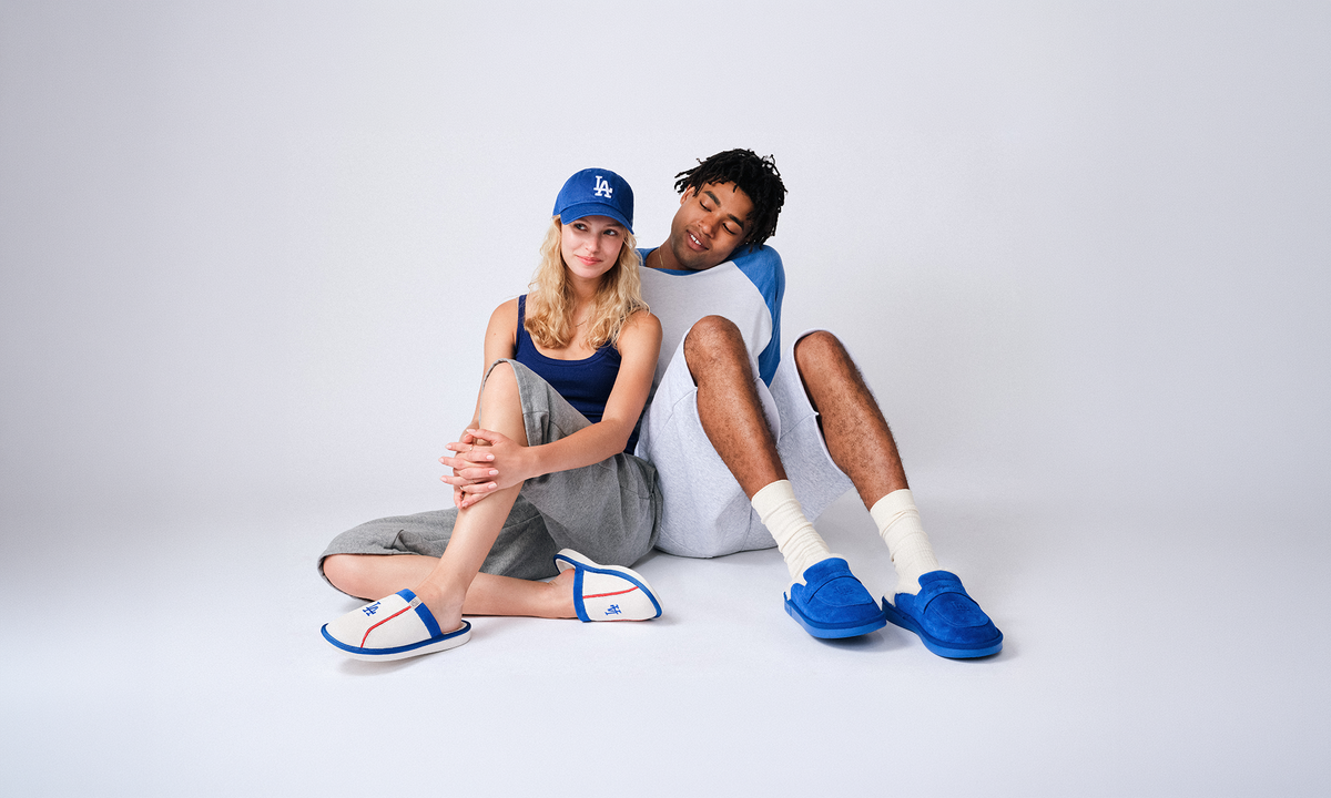 Two models sitting together on a light gray studio background. The woman wears a blue cap with an LA logo, a dark blue tank top, gray shorts, and blue-and-white sandals.
