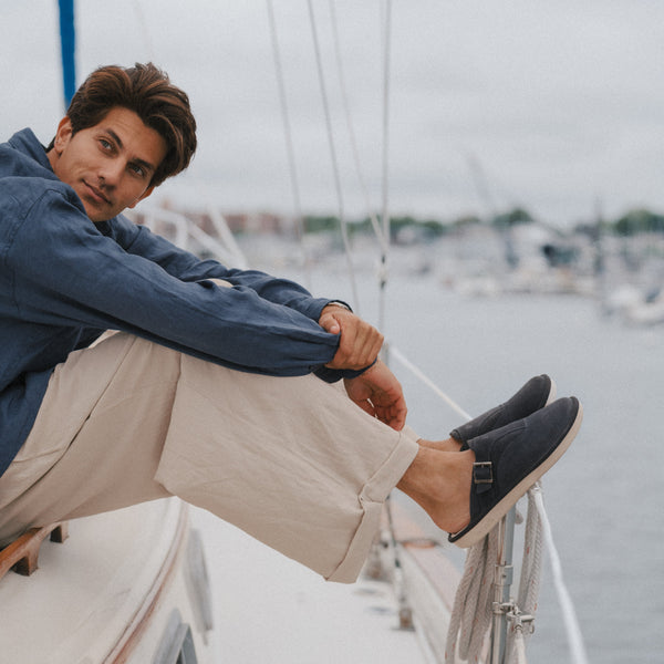 Man sitting on a sailboat rail, beige pants and charcoal slip-on shoes, looking over his shoulder toward a marina.