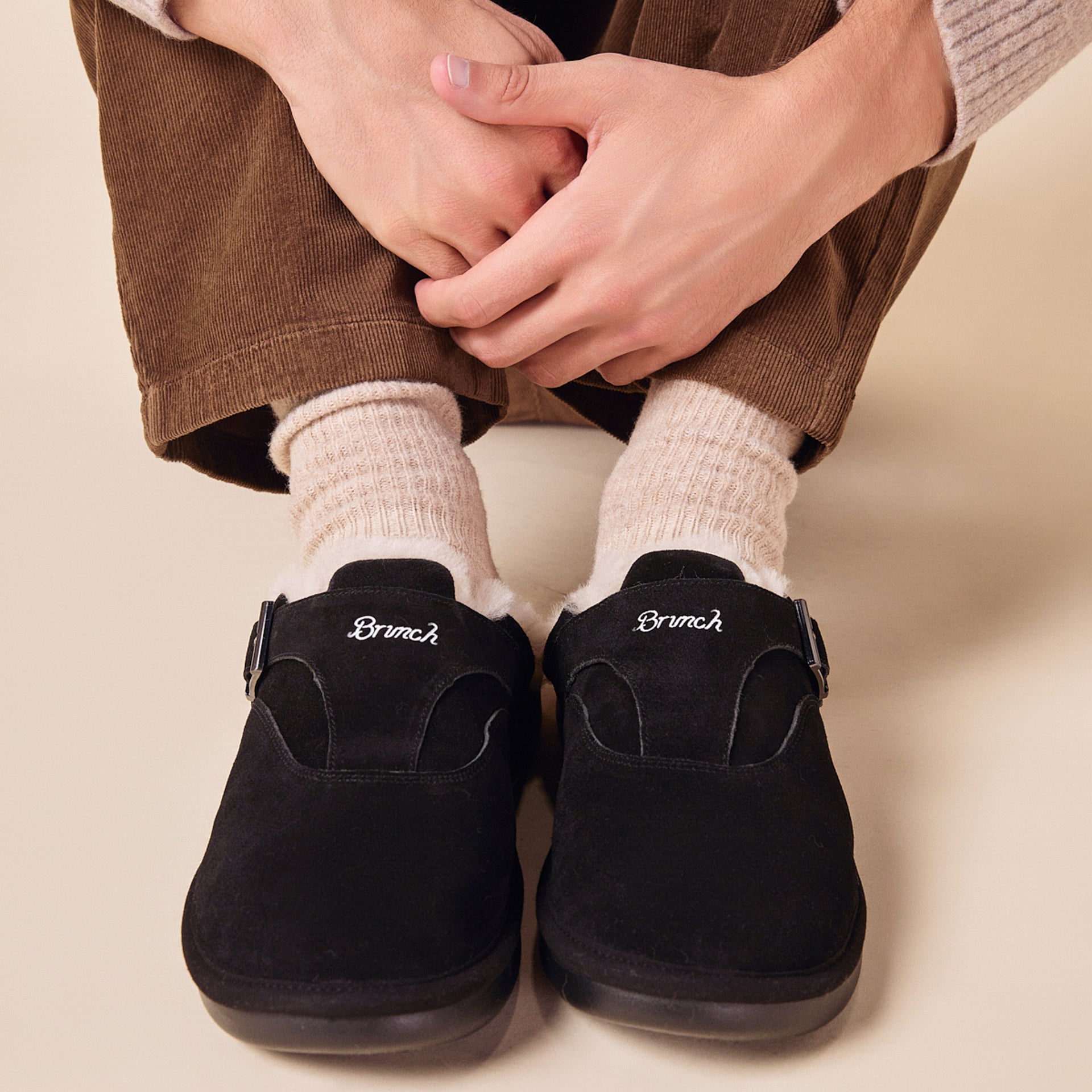 Seated person wearing beige socks and black suede slip-on clogs embroidered 'Brunch', hands clasped over knees.