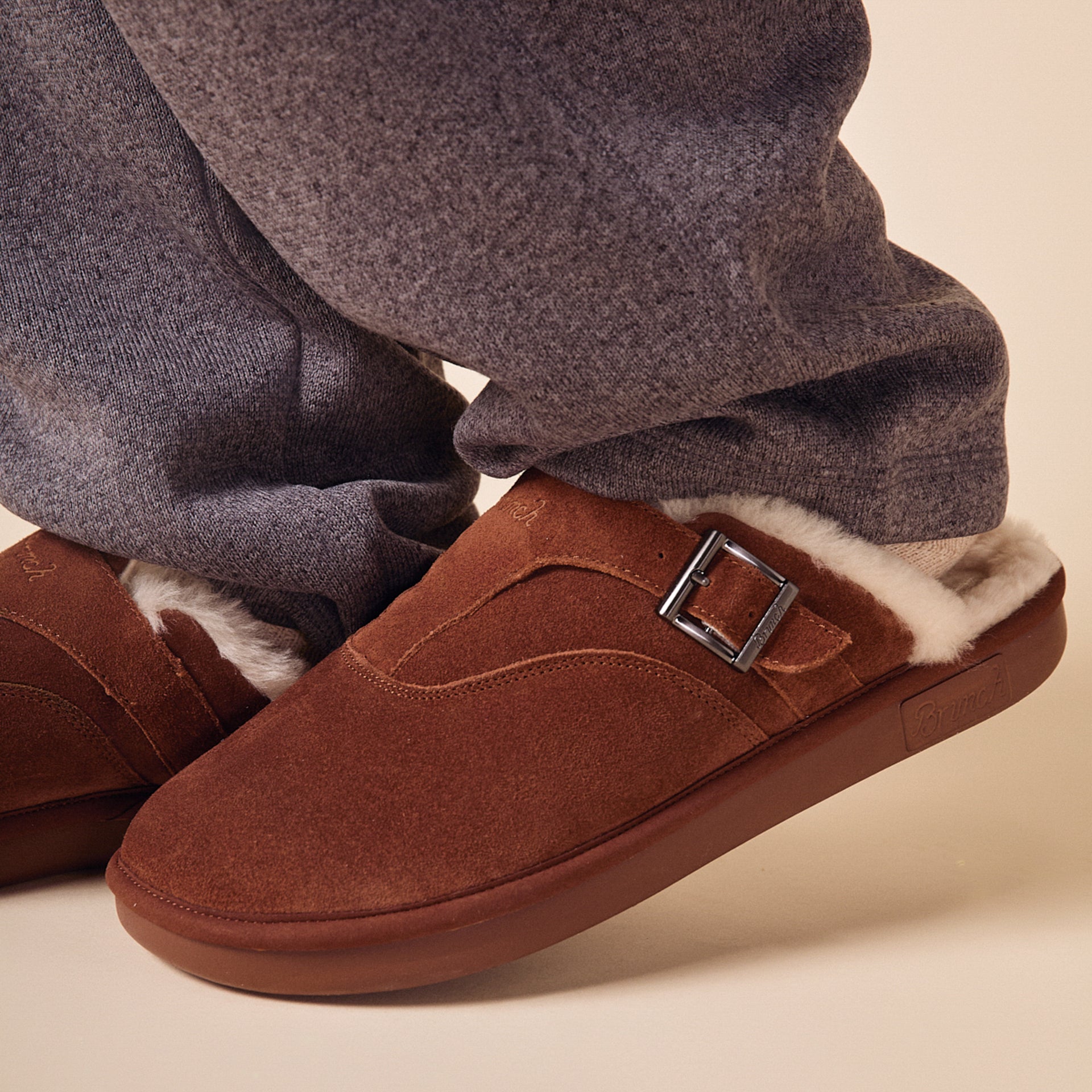 Brown suede shearling-lined slip-on clog with metal buckle, worn with gray sweatpants