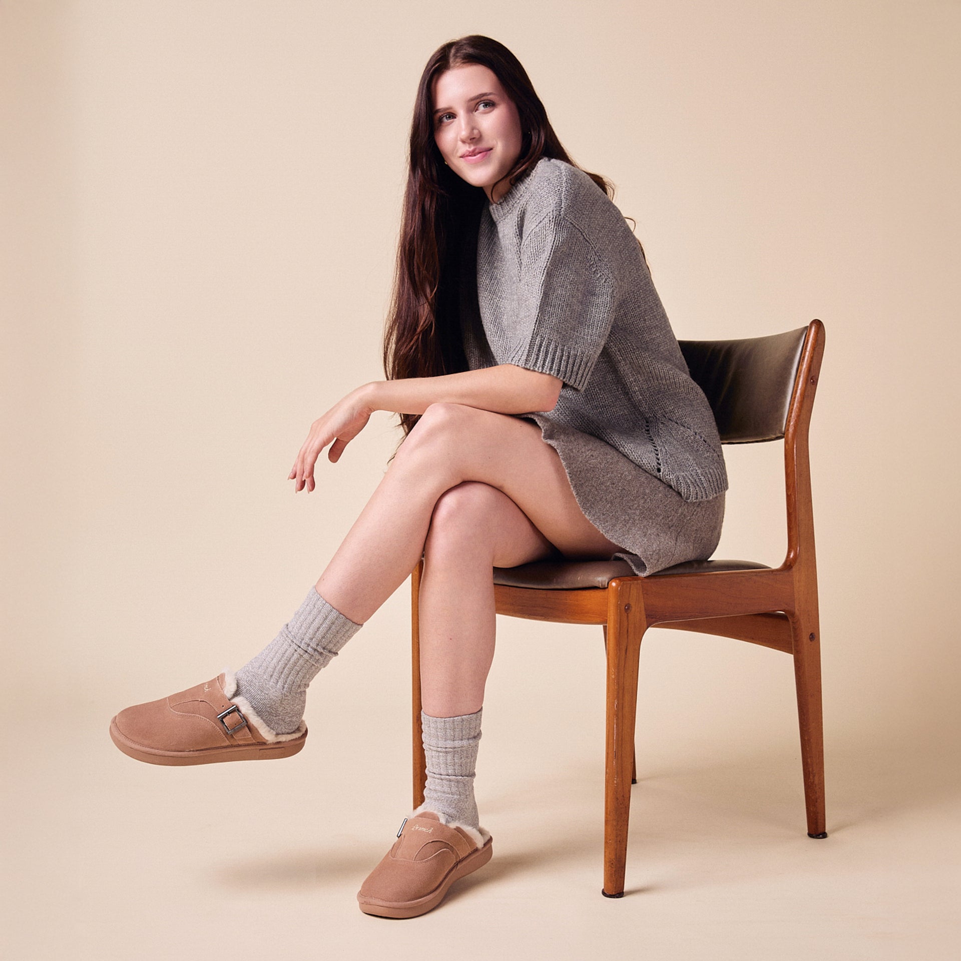 Woman seated on wooden chair wearing a gray knit dress, ribbed socks and taupe shearling-lined slip-on clogs.