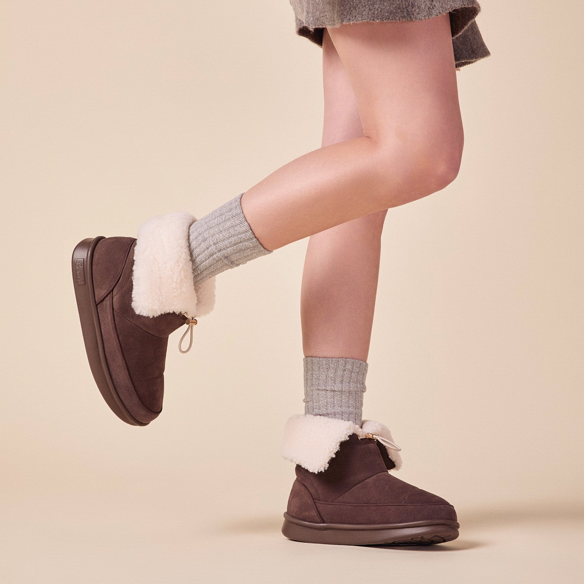 ankle-length brown suede shearling-lined slippers with folded cream cuff, worn with gray socks and a skirt, one foot lifted.