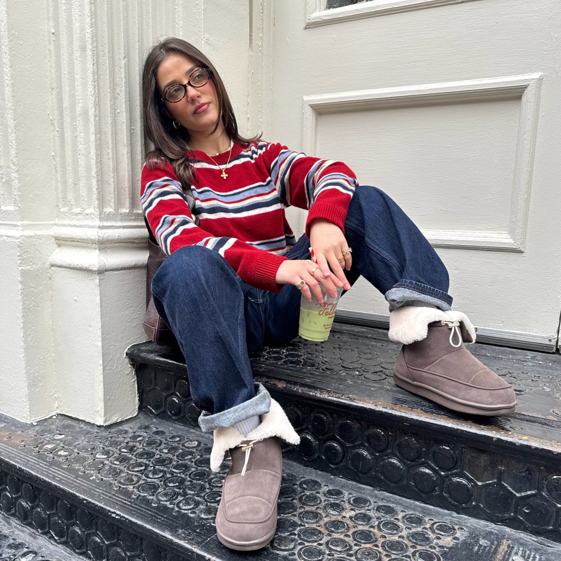 Woman in red striped sweater and glasses sitting on a stoop, holding a green drink, wearing cuffed jeans and brown shearling boots.