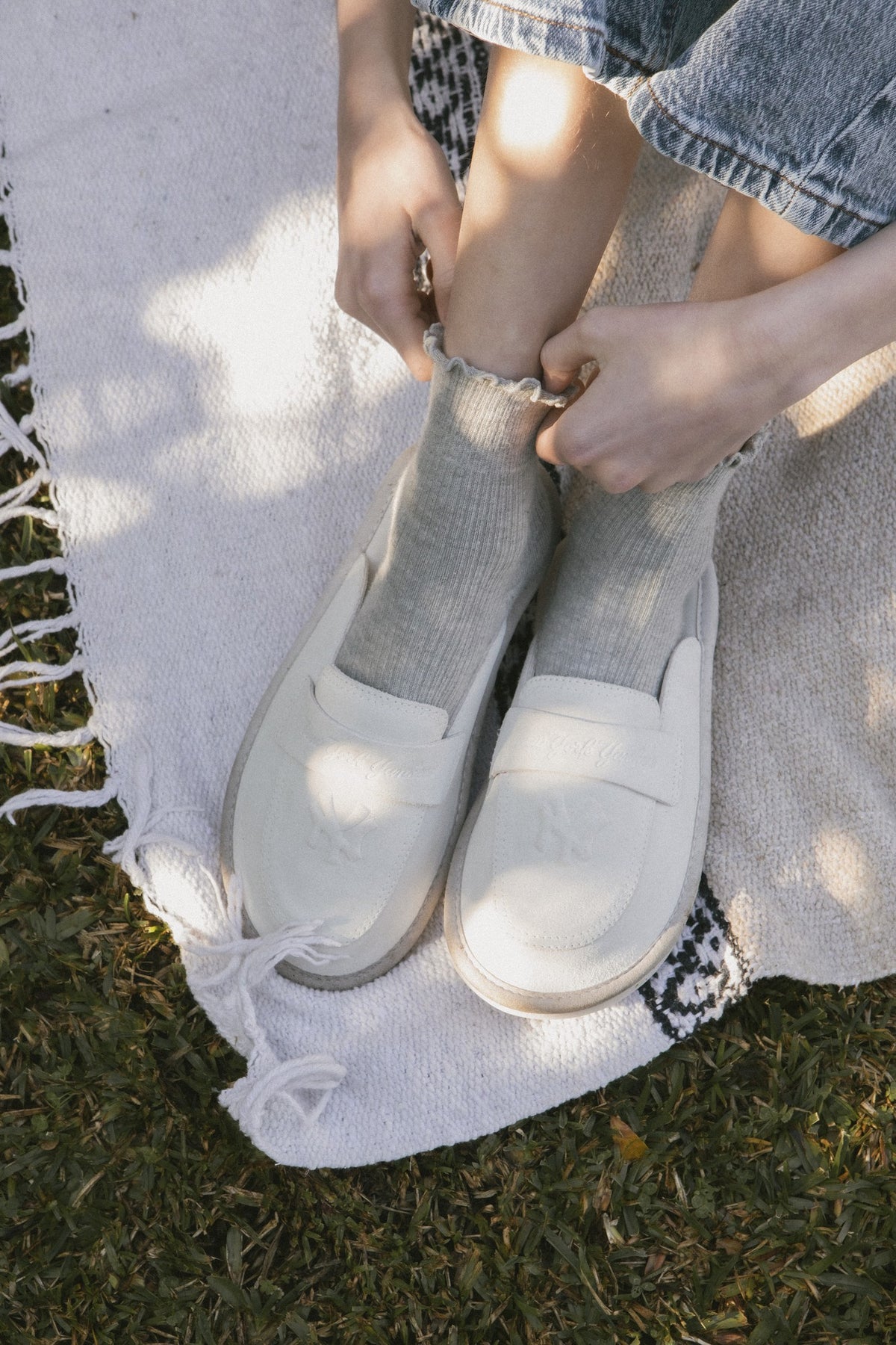 Top-down view of off-white suede loafers on feet showing embossed NY crest and penny strap, wearer adjusting socks on blanket
