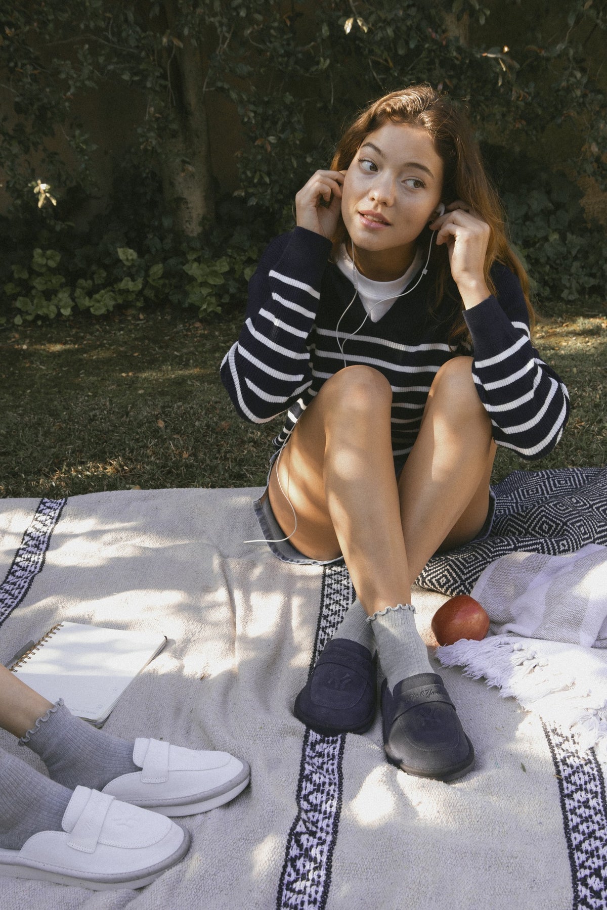Young woman on picnic blanket adjusting an earbud, wearing navy striped sweater and navy loafers