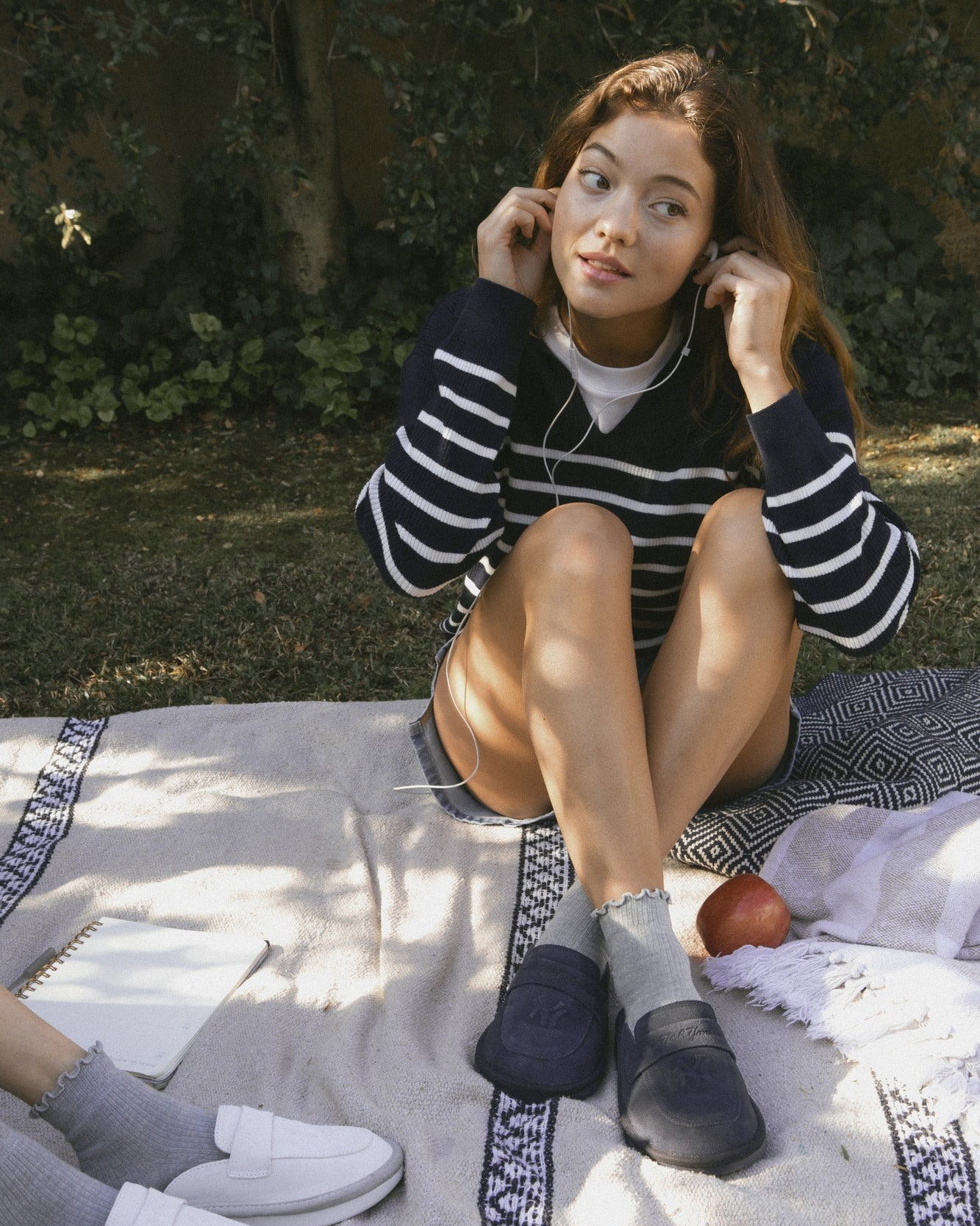 Woman sitting on a picnic blanket adjusting earphones, wearing a navy striped sweater and navy Yankees loafers