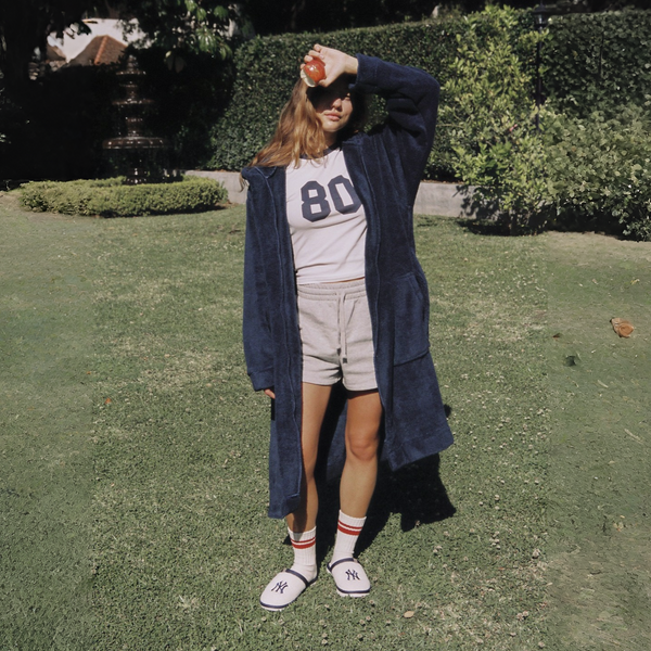 Person standing outdoors on grass wearing a long navy coat, white shirt with number 80, beige shorts, and white sneakers.